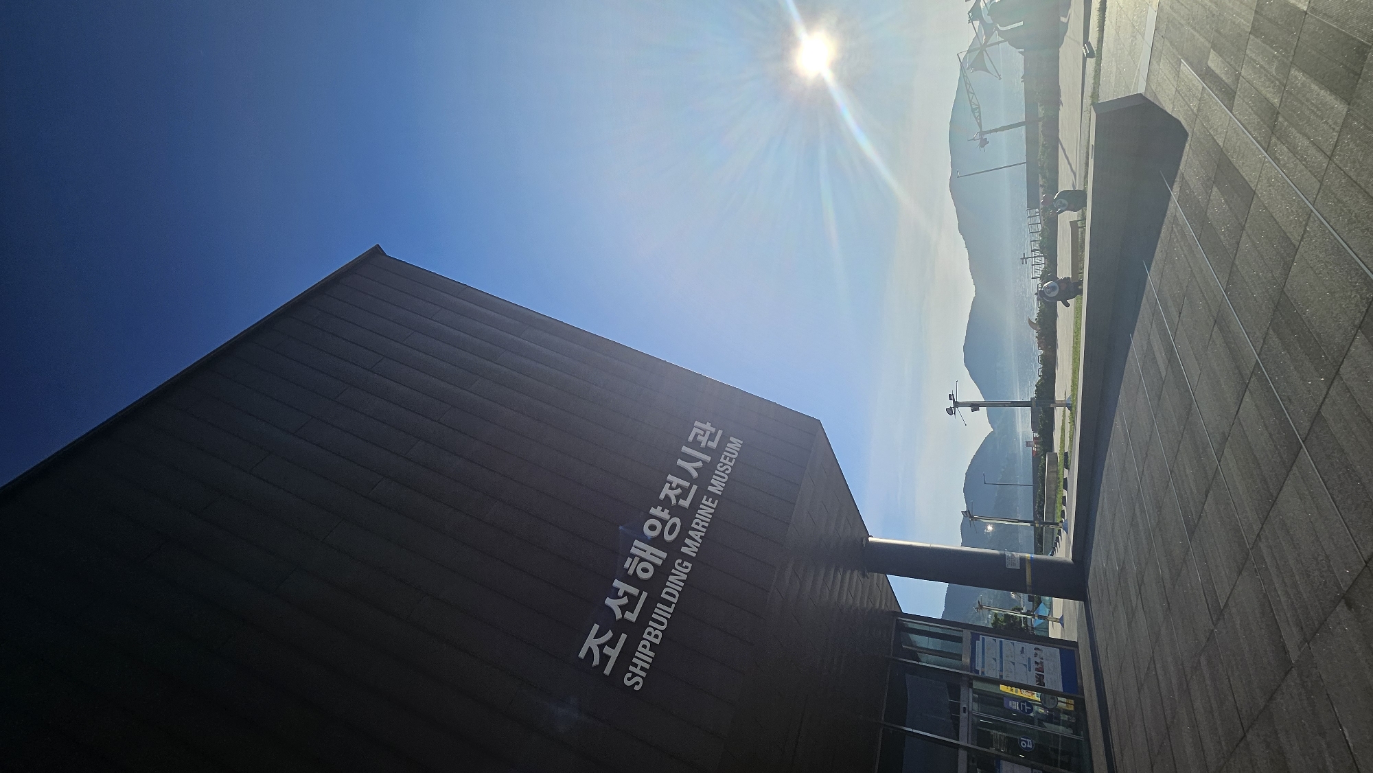 The image shows the exterior of the Shipbuilding Marine Museum in Geoje-si, South Korea, on a bright, sunny day. The museum's dark, angled facade, inscribed with Korean and English text reading "조선해양전시관 SHIPBUILDING MARINE MUSEUM," dominates the left side of the frame. The sky is a vibrant blue, with the sun visible in the upper right quadrant, casting a lens flare and rays of light across the scene.
In the background, a serene body of water is visible, bordered by misty, rolling hills. Along the waterfront, there are lampposts, a railing, and what appears to be a large crane structure on the right. In the middle ground, near a low, dark wall, two small, rounded, dark-colored figures resembling cartoon characters are seated. One appears to be looking towards the water, while the other is facing away from the camera. The foreground consists of a paved area with large, dark gray tiles. The entrance to the museum is partially visible on the far left, with glass doors and some informational posters displayed.