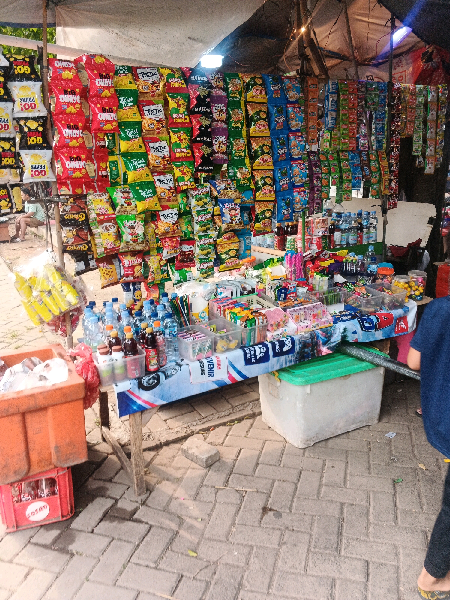 The image shows a street-side stall in Curug Kulon, Indonesia, selling a variety of snacks and drinks. The stall is covered by a tarp, and a variety of colorful snack bags are displayed vertically on racks. These include chips, cookies, and other packaged goods. Below the snacks, a table is laden with bottled drinks, including water bottles and what appear to be soft drinks and juices. There are also containers of candy and other small packaged items. To the left of the stall, two orange plastic crates are stacked, with a red crate containing bottles visible below. In the background, a person is seated on a stool, partially obscured. The stall is set up on a paved walkway, with cobblestone-like bricks forming the ground surface. The overall scene depicts a typical roadside vendor in Indonesia, offering a wide array of products to potential customers.