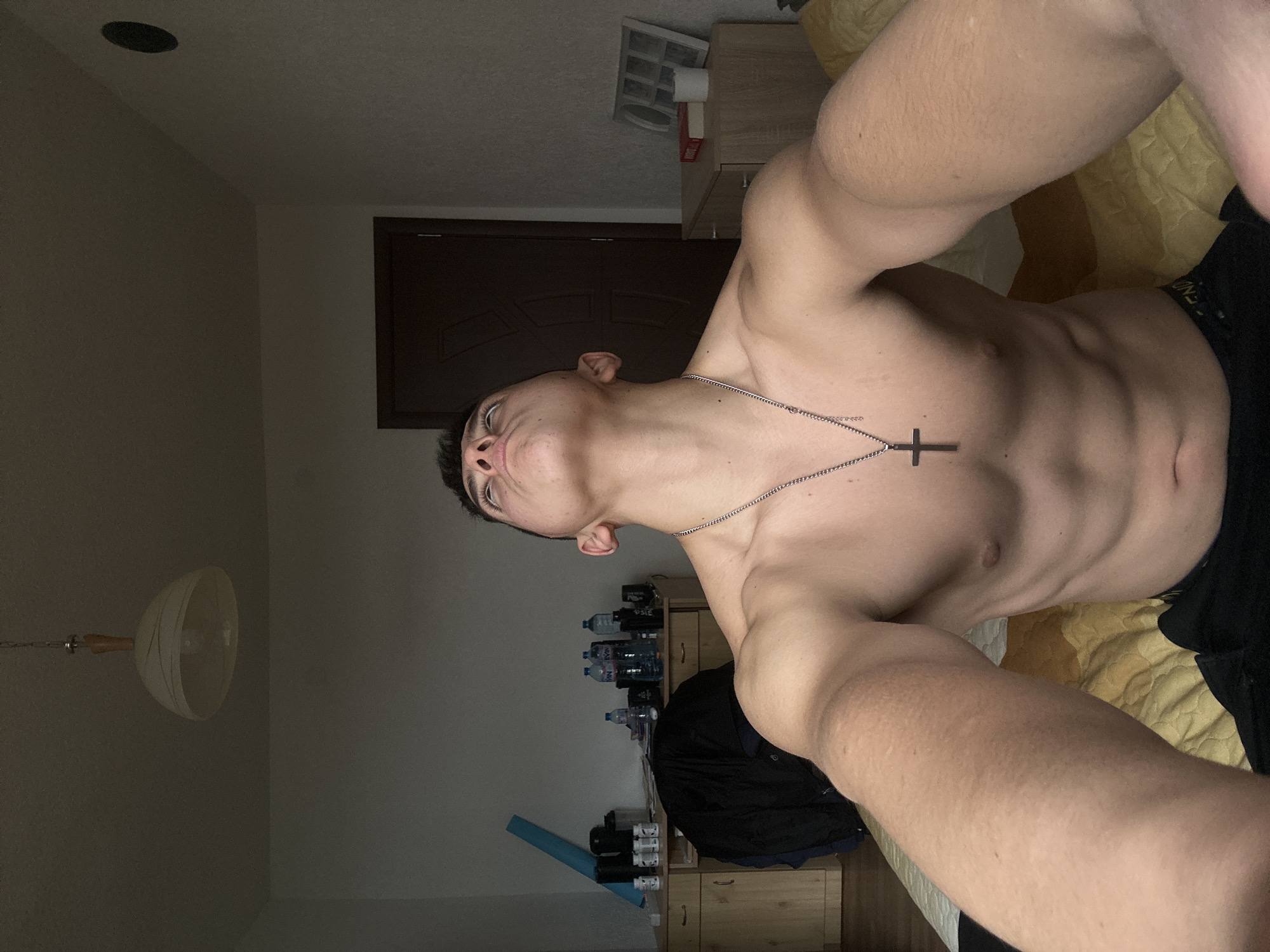 A shirtless man with a silver cross necklace is lying on a yellow and white bed. He is wearing black bottoms. The room contains furniture, including a wooden dresser with various items on it such as water bottles and workout supplements. There is a door in the background and a ceiling light fixture in the lower left. The image does not contain identifiable information to confirm that this location is in Zlatograd, Bulgaria.