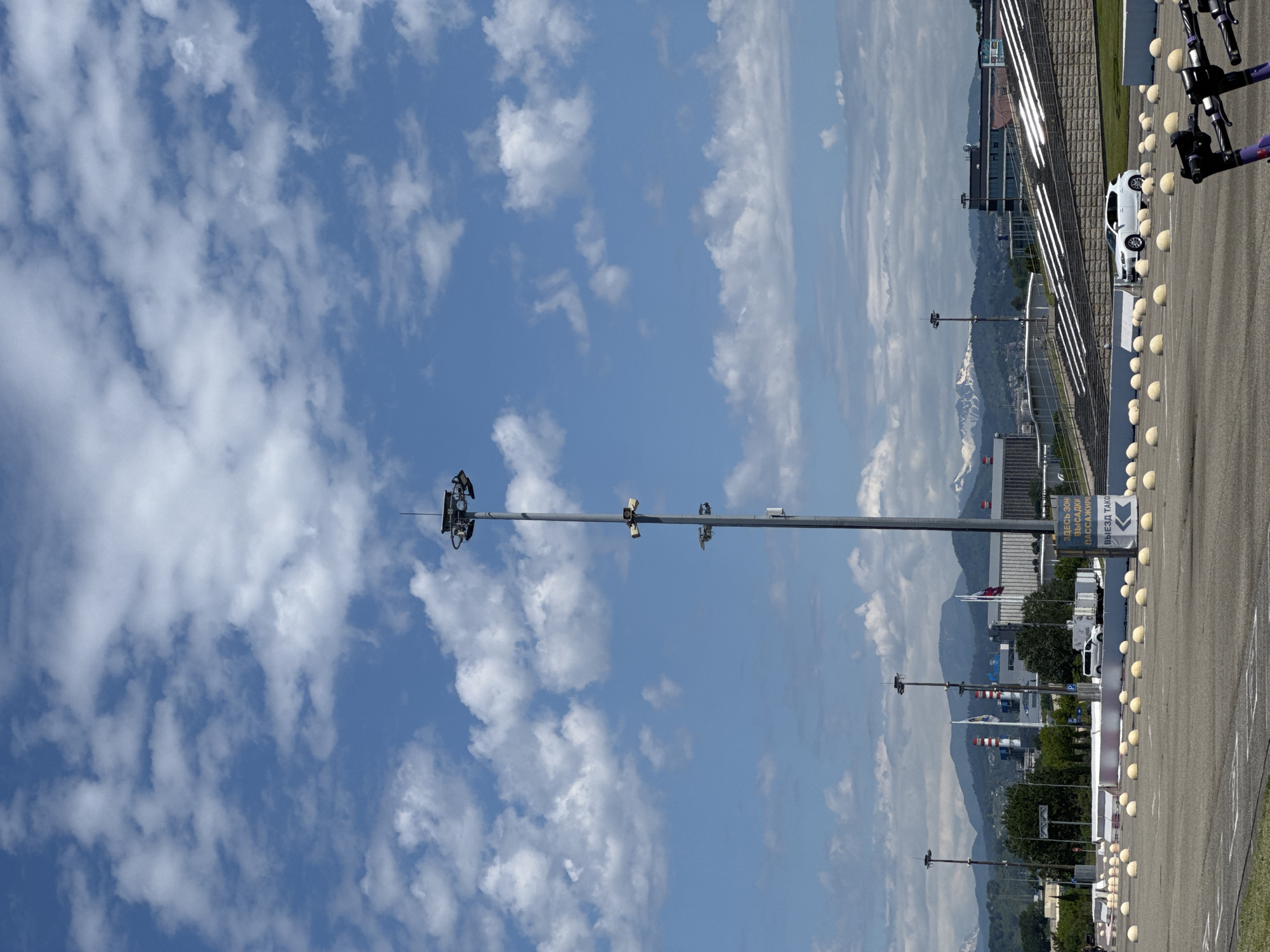 This outdoor image captures a wide panoramic view of a sunny day in Sirius, Russia, likely near a motor racing circuit or large event complex. The scene is dominated by a clear blue sky with scattered white cumulus clouds.
In the center of the frame, a tall, grey metal pole stands prominently, equipped with multiple cameras or surveillance devices. To the right, a wide paved area, possibly a road or part of a track, is visible, bordered by a long line of spherical, light-colored bollards. Behind these bollards, a white car is parked, and further to the right, several electric kick scooters are visible.
In the background, a majestic range of mountains stretches across the horizon, with some peaks still showing patches of snow, indicating their significant elevation. Various modern buildings are nestled between the paved area and the mountains.
A white sign, positioned near the bollards on the right, displays Cyrillic text. It reads: "ЗДЕСЬ ЗОН ВЫСАДКИ И/И ССАЖИР" (HERE IS THE DROPOFF/PASSENGER ZONE) and "ВЫЕЗД ТАКСИ" (TAXI EXIT), accompanied by a left-pointing arrow.
The time of day is clearly daytime, indicated by the bright lighting and the shadows. The weather is clear and pleasant, with good visibility. No people are visible, and no specific event or activity is taking place, presenting a serene view of the location.