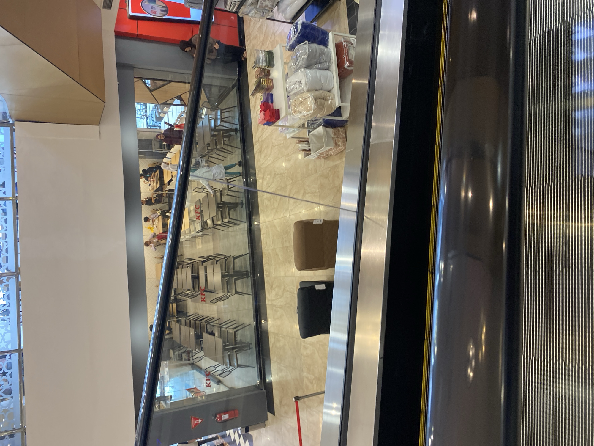 The image shows an indoor scene, likely a shopping mall in Banda Aceh, Indonesia, with an escalator in the foreground. Below the escalator, a KFC restaurant is visible with tables and chairs set up for dining. Several people are seated at the tables, and some are walking around. The restaurant has large glass windows. To the right of the KFC, there is a retail display with various items, including bedding or textiles. In the background, there are more levels of the mall with railings and some decorative elements. Two chairs, one black and one brown, are placed on the floor near the escalator.