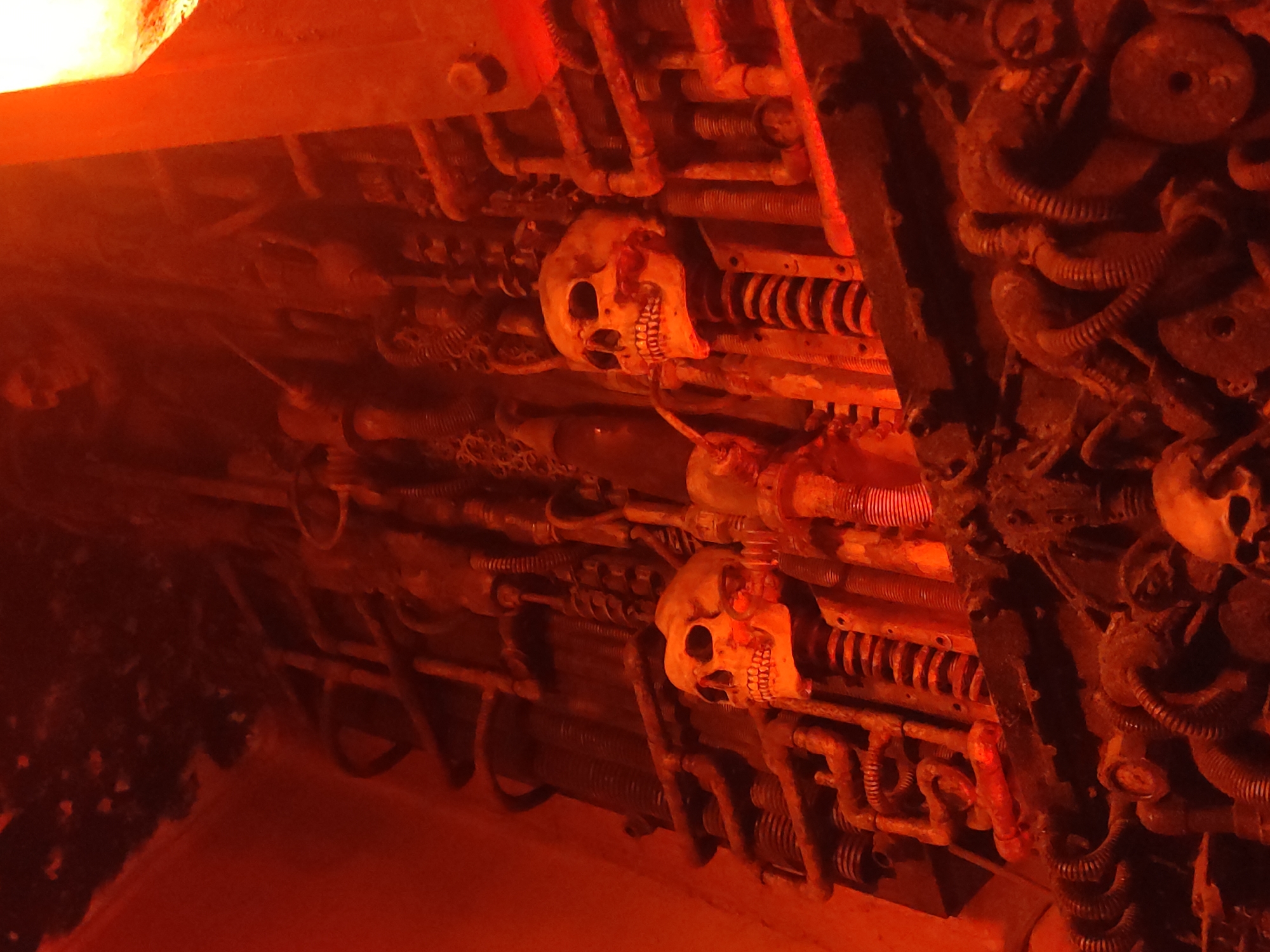 A close-up, medium shot captures a dimly lit, industrial-themed scene dominated by a warm, orangey-red glow, likely from artificial lighting. The focus is on a complex arrangement of metal pipes, coils, and mechanical components, creating a somewhat unsettling and cluttered aesthetic. Two human skulls are prominently displayed, seemingly integrated into the machinery. The skull on the left appears to be attached to a coiled spring mechanism. The skull on the right is also supported by a similar coiled structure. Both skulls are pale and feature distinct eye sockets and teeth, suggesting they are props or decorative elements. The background is filled with more pipes, wires, and metallic elements, some of which appear rusted or weathered. The overall impression is one of a macabre, possibly artistic, installation or a themed environment. The location is identified as Unknown, Unknown.