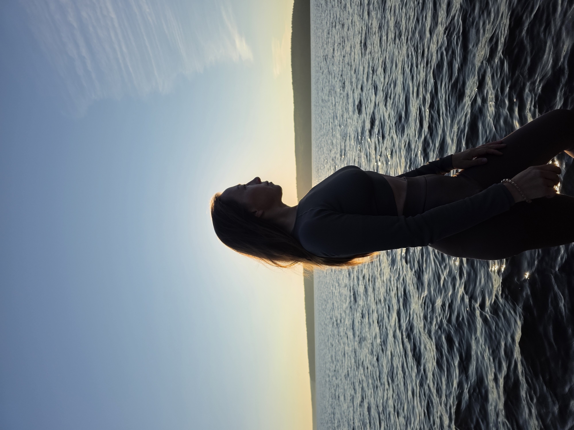 A woman stands in profile, facing right, against a twilight sky over a body of water. She is silhouetted by the setting sun, which creates a halo effect around her hair and the right side of her face. Her dark hair is long and falls past her shoulders. She is wearing a dark, form-fitting long-sleeved top that exposes her midriff and matching leggings. Her left arm is bent at the elbow, with her hand resting on her hip. Her right hand is relaxed at her side. A beaded bracelet is visible on her left wrist. The water in front of her is choppy, with ripples and glints of light reflecting off its surface. In the distance, a tree-lined horizon is visible, bathed in the warm glow of the sunset. The sky transitions from a pale yellow near the horizon to a clear blue overhead. The scene is peaceful and contemplative. The specific location within Chaikovsky, Russia is not discernable from the image alone.