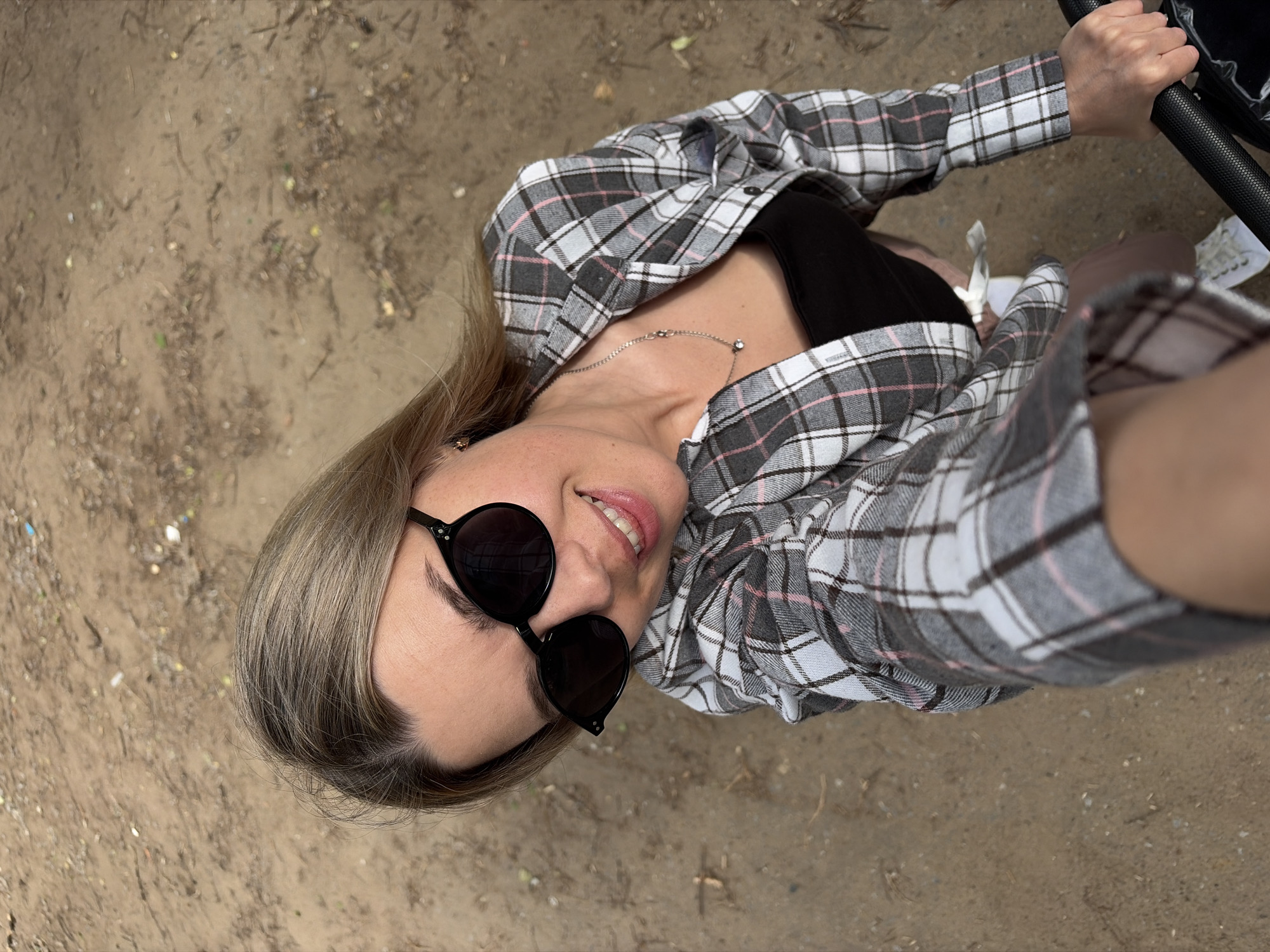 The image shows a woman taking a selfie outdoors, possibly in Chaikovsky, Russia, as indicated by the sandy ground with scattered leaves and twigs. She is wearing a black top, an unbuttoned grey and white plaid shirt with pink accents, and white sneakers. She has on black sunglasses and a silver necklace with two small pendants. Her hair is blonde and styled loosely. The woman is smiling and looking directly at the camera. In the lower right corner, a portion of a stroller handle is visible, suggesting she may be out with a child. The overall environment is natural and appears to be a park or wooded area.