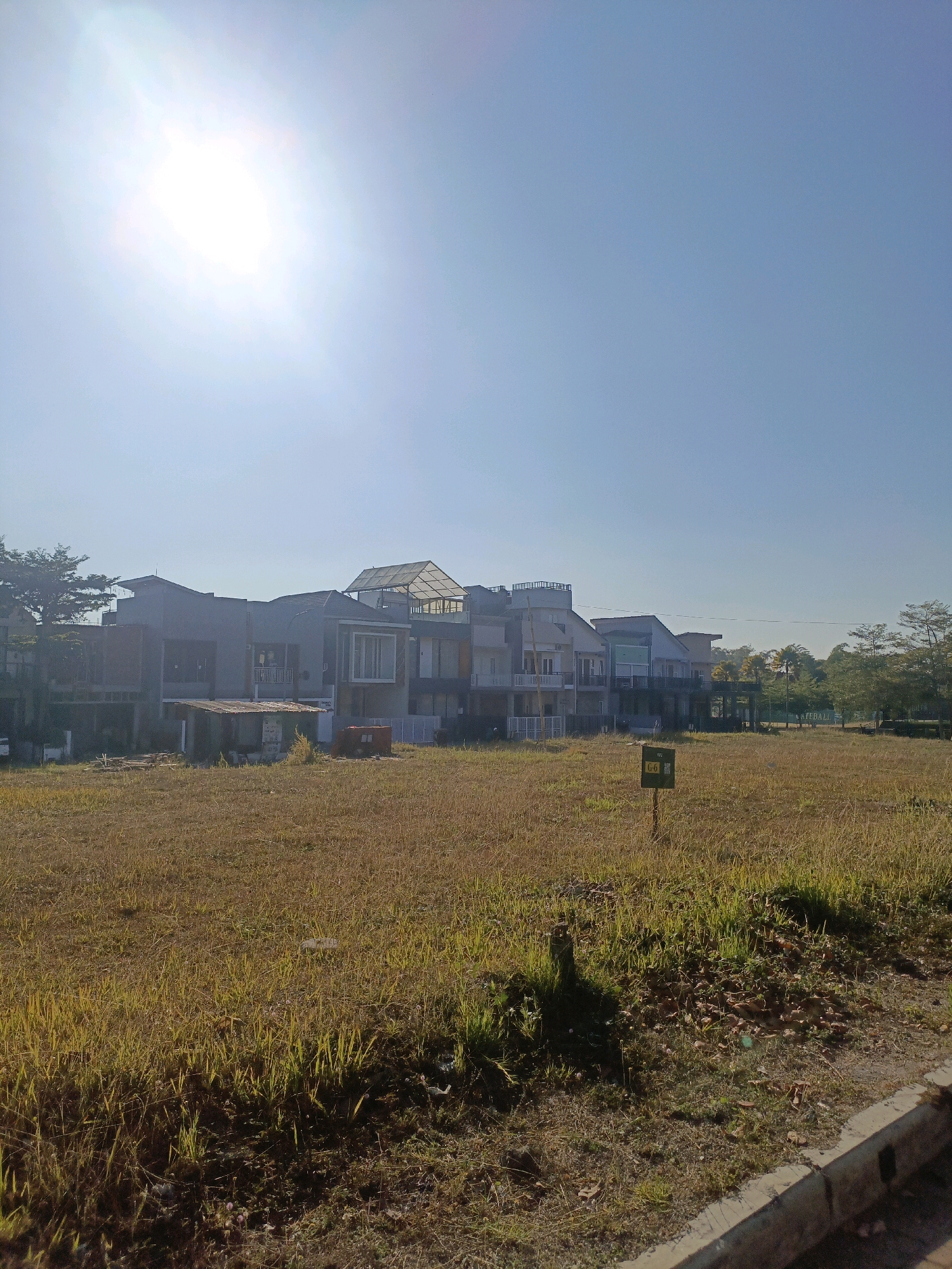 The image depicts a sunny day in Batu, Indonesia, with a clear blue sky. In the foreground, a field of dry, yellow grass is visible, with some green patches and scattered dry leaves. A concrete curb with a drainage grate runs along the bottom right edge of the image. In the middle ground, a small sign with the inscription "G-6" is planted in the grass. Behind the field, a row of modern, multi-story houses is under construction or recently built. These houses feature a mix of concrete and glass facades, with balconies and some having rooftop structures. A large, bright sun is prominent in the upper left corner, casting a lens flare across the sky. The overall scene suggests a developing residential area in a tropical climate.