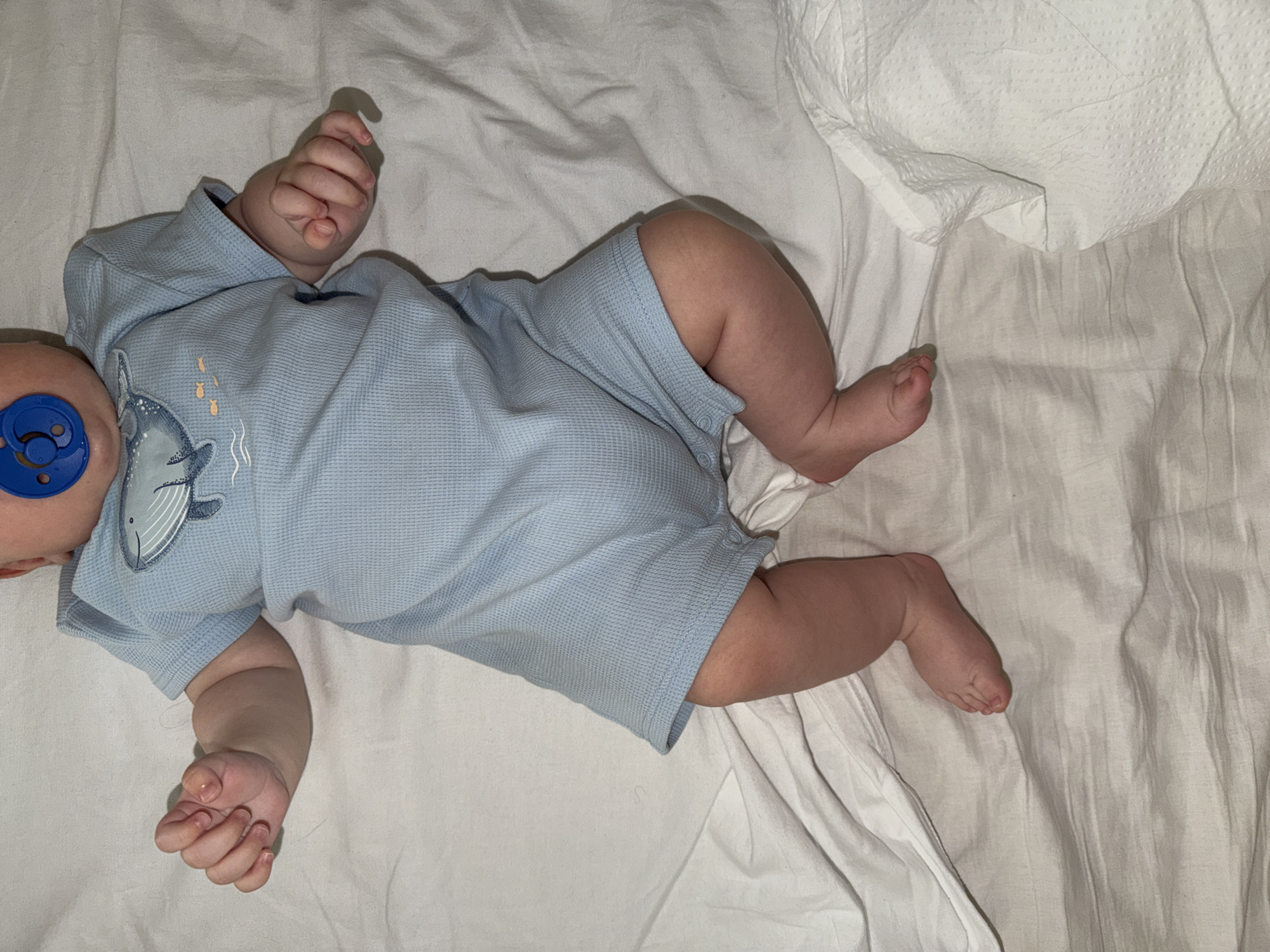 A baby is depicted lying on a white, textured sheet, likely resting or sleeping. The baby is wearing a light blue, waffle-knit romper with short sleeves and legs, adorned with an illustration of a whale and small fish on the chest area. A blue pacifier is visible in the baby's mouth, with only the side of the baby's face and ear visible. The baby's arms are bent with hands loosely fisted, and legs are slightly bent. One arm is positioned near the head, and the other along the side. The lighting is somewhat dim, casting soft shadows, suggesting an indoor setting, possibly during the evening or night. A crumpled piece of white fabric, perhaps a blanket or towel, is visible in the upper right corner. No text is visible in the image. The scene conveys a sense of peacefulness. This media file was captured in Khimki, Russia.