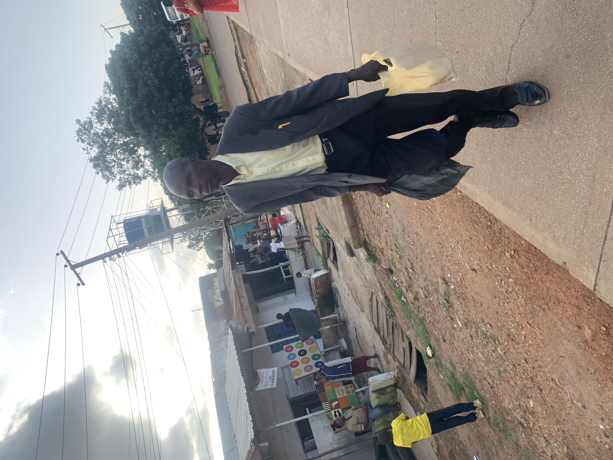 A man in a dark suit and a light yellow shirt, holding a yellow plastic bag, walks on a paved surface. He is wearing black shoes and a belt. To his right, a dark bag is partially visible on the ground. In the background, there are buildings, trees, and a cloudy sky. Electrical wires are prominent across the scene. Further back, other people are visible, some standing, some sitting. A building with a blue and green sign is visible on a metal structure. Another building appears to be a shop with merchandise displayed, including colorful items on the wall and on the ground. A sign on the shop reads "Nkasi House" and "call: 014092840". The scene appears to be an outdoor marketplace or street in a populated area. The lighting suggests it is daytime, possibly late afternoon, with the sun partially obscured by clouds. The ground is a mix of paved areas and dirt.