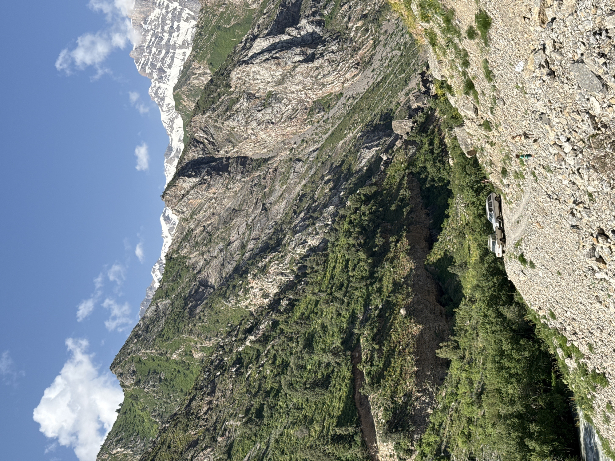 The image depicts a mountainous landscape with a clear blue sky dotted with white clouds. The mountains are rugged and steep, with some peaks covered in snow. Lush green vegetation covers the lower slopes, giving way to rocky terrain higher up. A dirt road winds through the mountains, and several vehicles, including SUVs, are parked on the side of the road. A few people are visible on or near the road. The scene suggests a remote, natural environment, possibly a travel destination or a hiking area. The location is identified as an unspecified city in Uzbekistan.