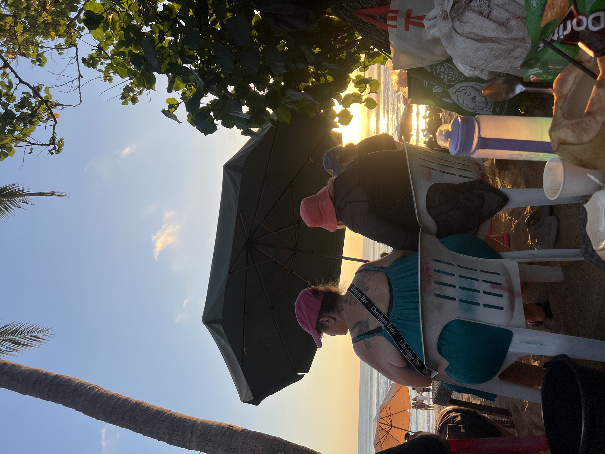 The image depicts a beach scene in Kuta, Indonesia, during sunset. Three people are seated on white plastic chairs facing the ocean. The person in the foreground, on the left, is a woman wearing a teal dress and a pink baseball cap. She has multiple tattoos on her back and is wearing a black crossbody bag with "Christian Dior" written on it. To her right, another person wears a pink bucket hat and a dark jacket. A third person is partially visible behind them, wearing a blue cap.
In the foreground, several objects are placed on the sand, including a black bucket, a white plastic cup, a purple and white water bottle, a metal spoon, a half-coconut with a straw, a bag of chips, and other miscellaneous items. A large dark green umbrella is positioned over the seated individuals, and a bright orange umbrella is visible further down the beach. The sun is setting over the ocean, casting a warm glow on the scene. Palm trees and foliage frame the right side of the image. People are visible walking on the beach in the distance.