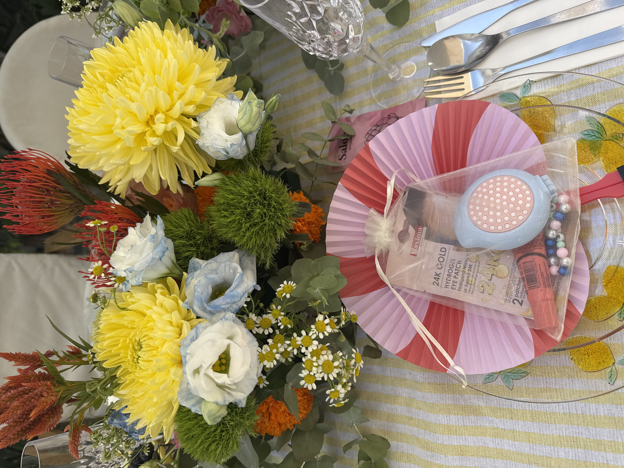 The image captures an overhead view of a set table, likely for an outdoor event in Agios Tychona Community, Cyprus, given the presence of a bouquet of flowers and table setting. A floral arrangement dominates the upper portion of the frame, featuring bright yellow chrysanthemums, delicate blue and white roses, fiery red pincushions and celosia, and small white daisies. The flowers are interspersed with greenery.
In the foreground, a place setting includes a crystal wine glass, silverware (a fork, spoon, and knife), and a clear plate decorated with yellow lemon slices. Centered on the plate is a folded red and pink fan, within which is a small gift bag. The bag contains beauty products, including what appears to be a tube of lipstick and a compact with a pink patterned lid, alongside a sample of 24K GOLD Hydrogel Eye Patch. The table is covered with a yellow and white striped tablecloth. The lighting suggests it is daytime, possibly a garden or patio setting. There are no people visible in the image.