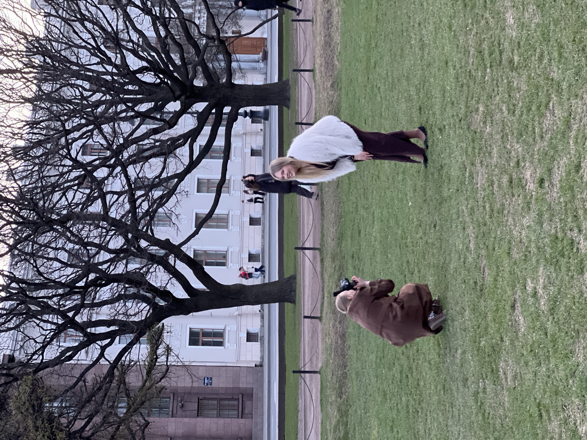 The image shows a woman being photographed on a grassy lawn in Saint Petersburg, Russia. The woman is wearing a white fur coat and a maroon dress. She is standing in front of a large, bare tree and a light-colored building with windows. A photographer in a brown coat is kneeling on the grass, taking her picture. A fence runs parallel to the building. There are other people walking in the background, near the building and trees. The scene appears to be outdoors, during the day.