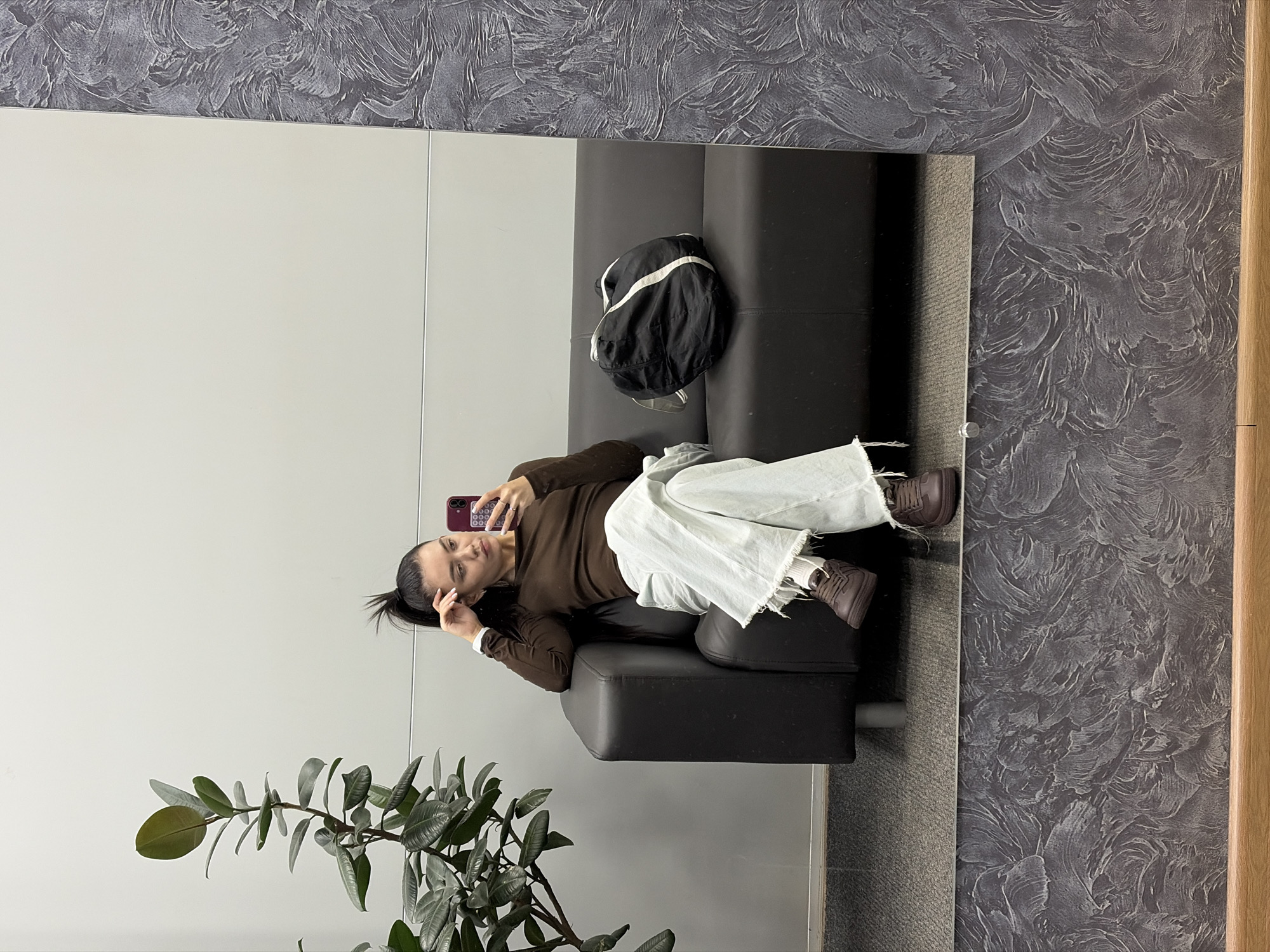 A mirrored view depicts a woman reclining on a black couch. She is wearing a brown long-sleeve shirt, white pants, and brown sneakers, holding a smartphone up as if taking a selfie. A black and white bag sits near the top of the couch. To the left, a potted plant is visible. The mirror is positioned against a wall with a textured gray pattern, bordered by a light-colored wall to its left and a wooden strip to the right. The location is Chaikovsky, Russia.