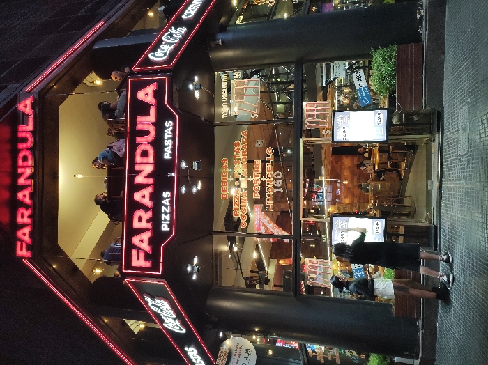 The image depicts the nighttime exterior of the "FARANDULA PIZZAS PASTAS" restaurant, located on Corrientes Avenue in Buenos Aires, Argentina. The two-story building features prominent red neon "FARANDULA" signs on both its upper and lower levels. Below the lower "FARANDULA" sign, "PIZZAS" and "PASTAS" are displayed in white neon, and "Coca-Cola" logos are integrated into the red neon border.
The ground floor features a large glass facade, revealing an illuminated interior with multiple patrons seated at tables. Spanish text on the windows advertises "BEBIDAS," "PIZZA + PASTAS COCINA ELABORADA," "POSTRE," and "LIMONCELLO 160," along with various prices, including "1.999" and "2.999." The upper floor is an open-air balcony dining area where at least four individuals are seated at tables under suspended lighting.
On the light-colored, patterned tiled sidewalk outside the ground floor entrance, two individuals are observing an illuminated digital menu display. The person on the left wears a dark long-sleeved top, white shorts, and dark sneakers. The person on the right, dressed in a dark t-shirt, dark shorts, and dark sneakers with white soles, appears to be interacting with the screen. Dark architectural columns frame the entrance, and planters containing green foliage are positioned at the building's base.