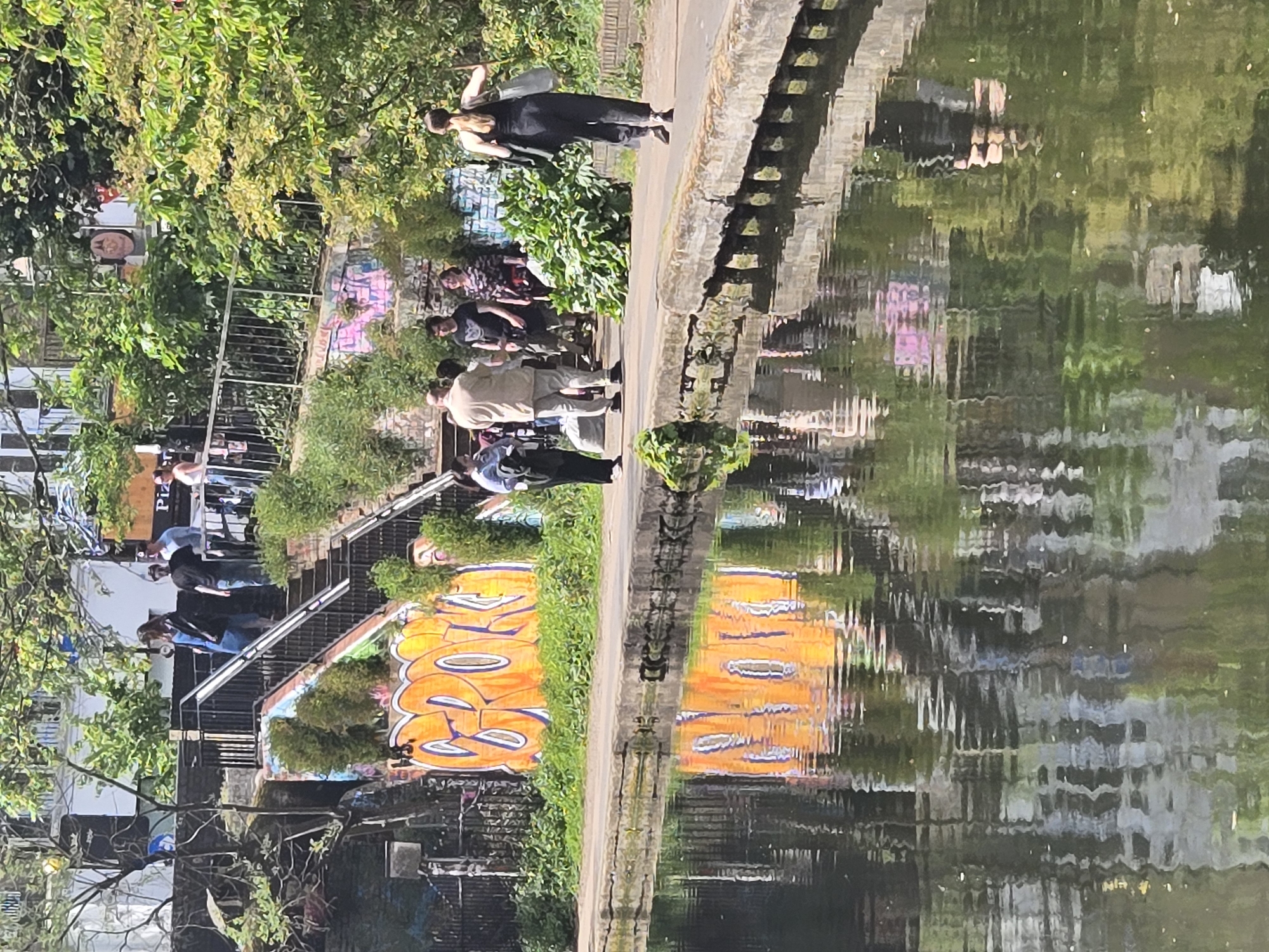 The image shows a canal scene in London, United Kingdom. A group of people stands along the canal path near a set of stairs leading to a higher level. Graffiti art is visible on walls near the stairs. One person is walking away from the group. The water reflects the scene, creating a distorted mirror image. The environment includes greenery such as trees and grass along the canal.