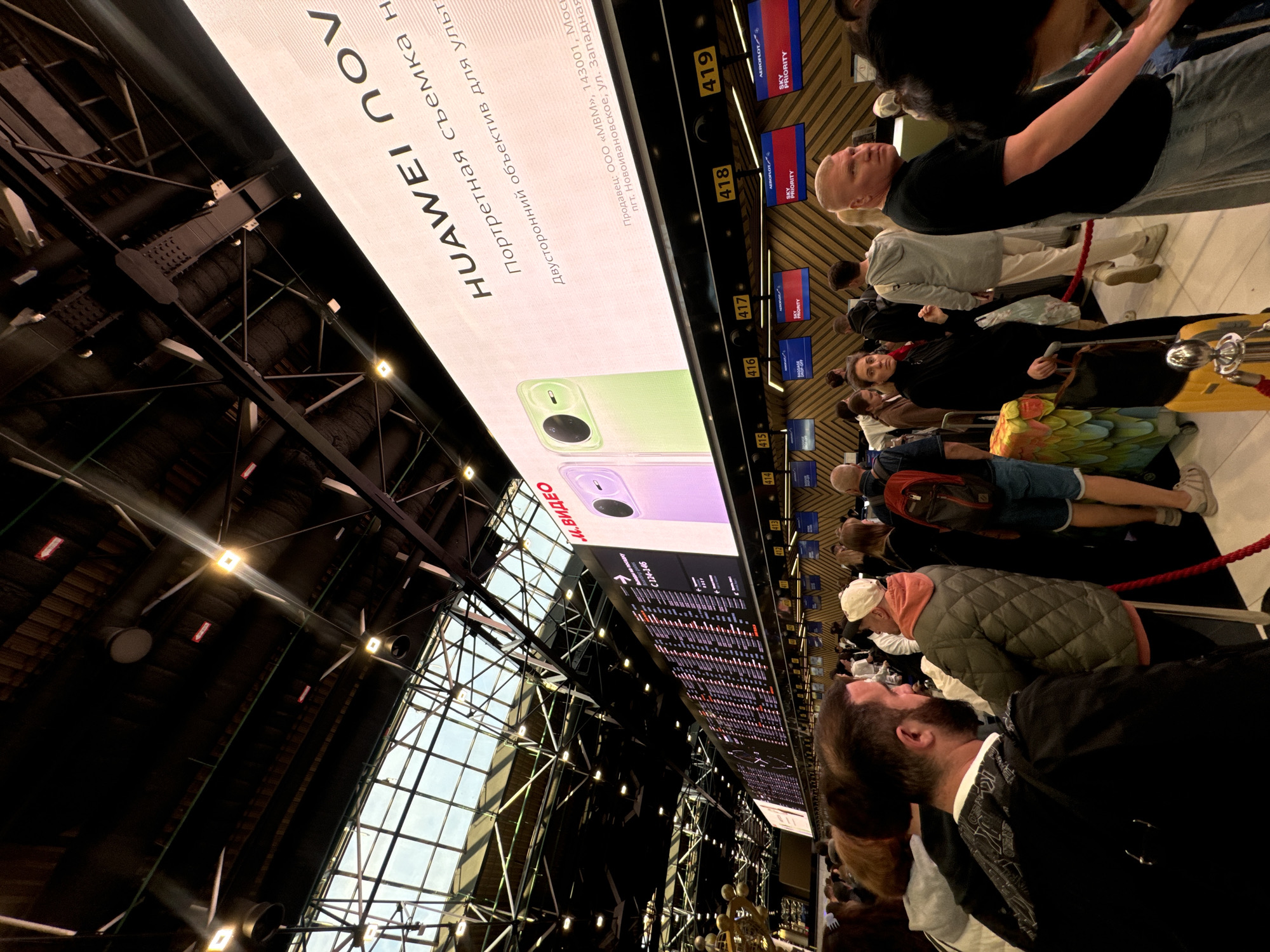 This image depicts a scene inside what appears to be an airport in Khimki, Russia, judging by the Cyrillic text and the presence of flight information displays. A large digital screen shows a Huawei advertisement with Cyrillic text reading "HUAWEI NOV" and marketing phrases in Russian, suggesting the phone's portrait shooting capabilities and a dual-sided lens. Below the advertisement is a flight information display board with departure or arrival times. Several people stand in line at check-in counters, likely waiting for their flights. The counters are numbered from 415 to 419 and display airline logos and "SKY PRIORITY." The overall lighting is bright, and the interior architecture features metal beams and a glass roof.