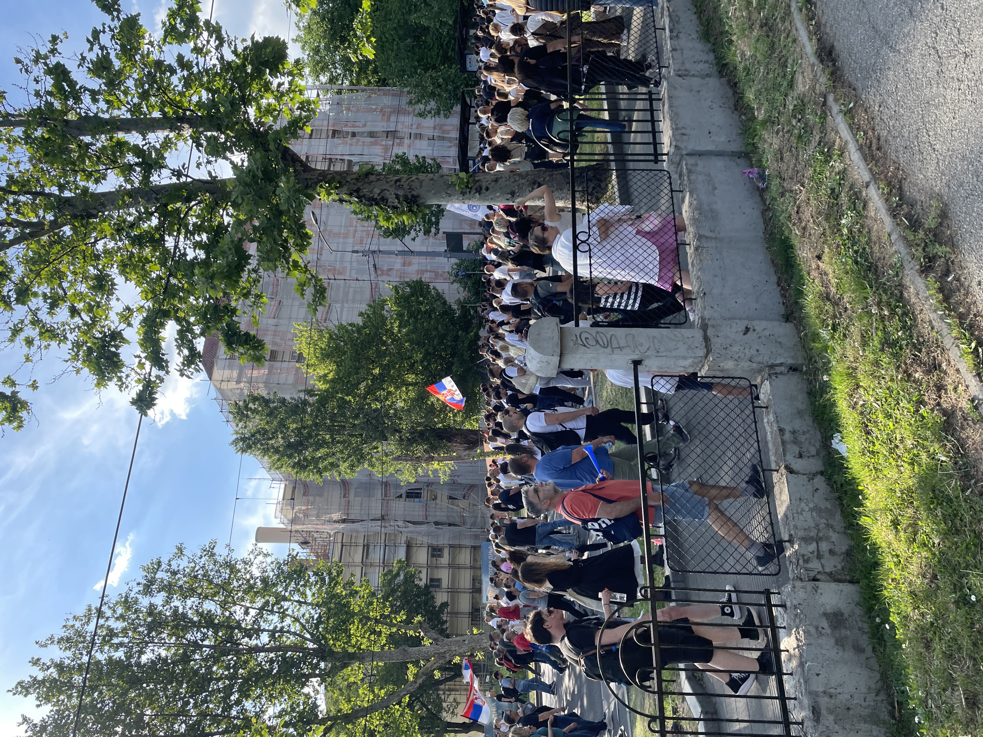 A crowd of people is gathered in Vracar Urban Municipality, Serbia, along a street bordered by a black metal fence and a concrete ledge, with a grassy area and paved road to the right. Many people are wearing light-colored clothing and some are holding flags of Serbia. A large building under construction is visible in the background, partially obscured by trees. The sky is blue with some clouds. The image suggests an outdoor event or gathering along a city street.