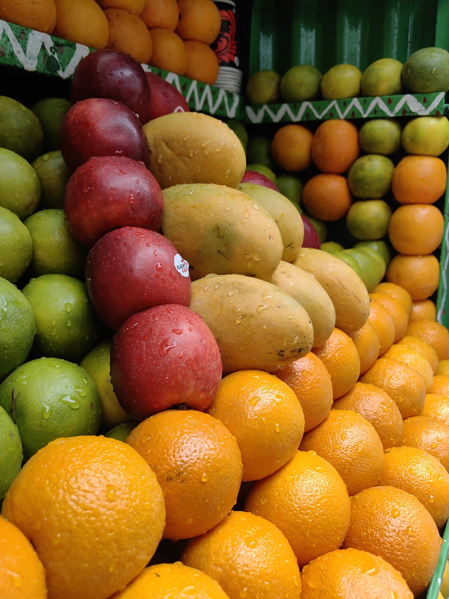 The image depicts a vibrant display of fresh fruits, likely at an outdoor market stall in Vashisht, India. A variety of fruits are artfully arranged in tiered stacks. In the foreground and midground, bright orange fruits, possibly oranges or tangerines, are piled high, their textured skin glistening with water droplets. Behind these, a row of smooth, yellow fruits, which appear to be mangoes, are visible. Interspersed among the mangoes and in front of them are deep red apples, also speckled with water. Further back, a selection of green fruits, likely limes or green apples, are stacked in rows. The fruits are presented on green shelving units, with some decorative elements on the edge of the shelves. The overall impression is one of abundance and freshness, characteristic of a local fruit vendor in Vashisht. There are no people or animals visible in the frame. The environment appears to be outdoors, with the focus solely on the meticulously arranged fruit stall.