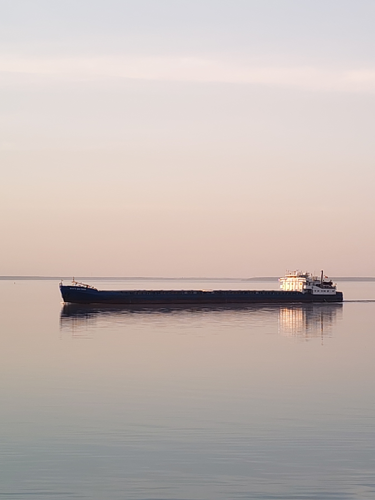 The image depicts a long, dark-colored cargo ship moving across a calm body of water under a pale sky. The lighting suggests either dawn or dusk. The ship's structure is visible, including the front section and a larger, brighter structure at the rear where light reflects off its surfaces. The water reflects the ship and the surrounding light, creating a mirror-like effect. The location is Gorokhovo, Russia, based on the title of the image.