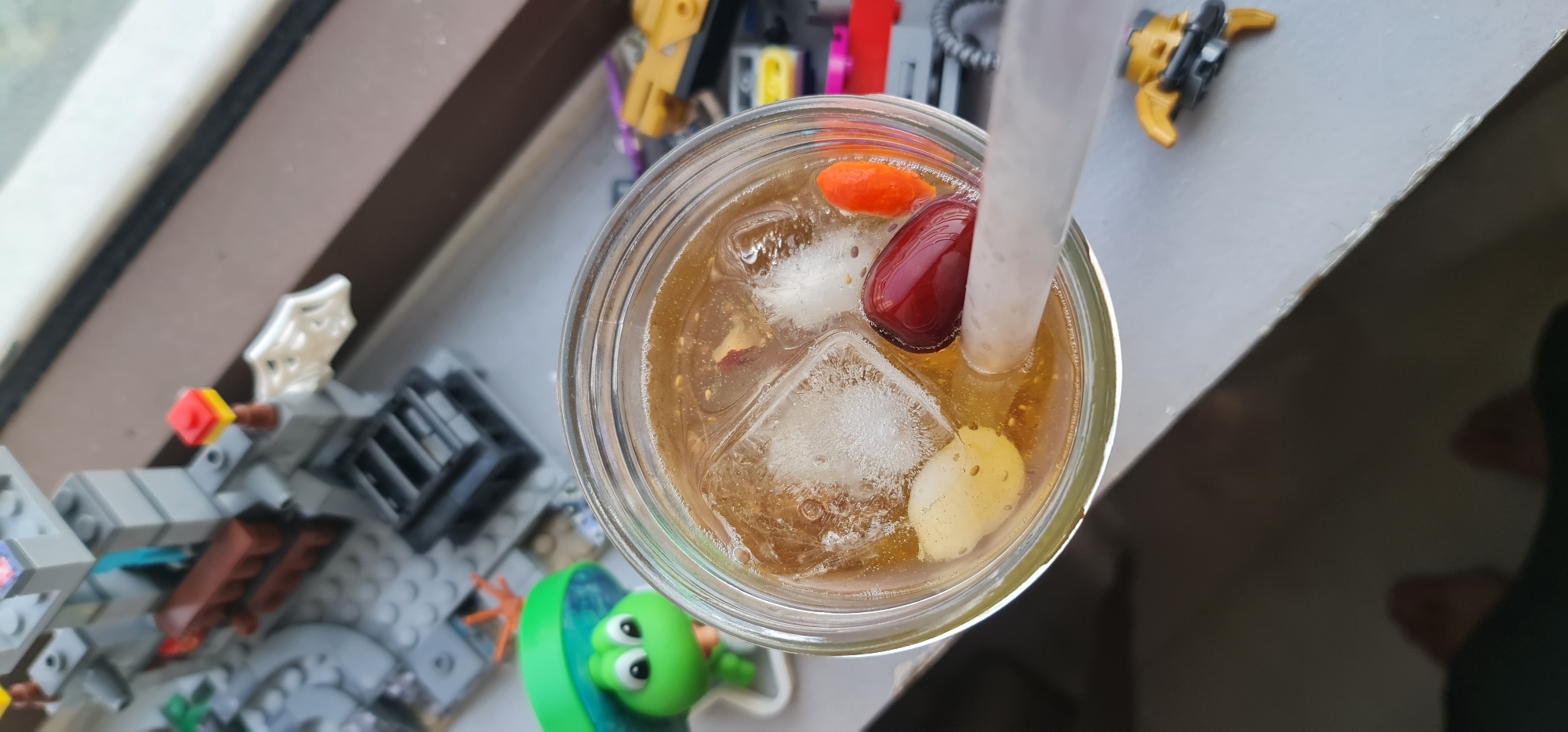 A top-down shot shows a glass of iced herbal tea on a white surface with various objects around it. The tea contains ice cubes, a white, bulbous fruit, a reddish-brown fruit, and an orange fruit. A white straw is inserted into the drink. Surrounding the glass are Lego toys: a gray castle structure, a yellow and black figure, and a green alien-like character. The location is likely in Đà Nẵng, Vietnam, based on the presence of the herbal tea, a popular beverage in the region. The background is partially obscured, but a window frame is visible at the top of the image.