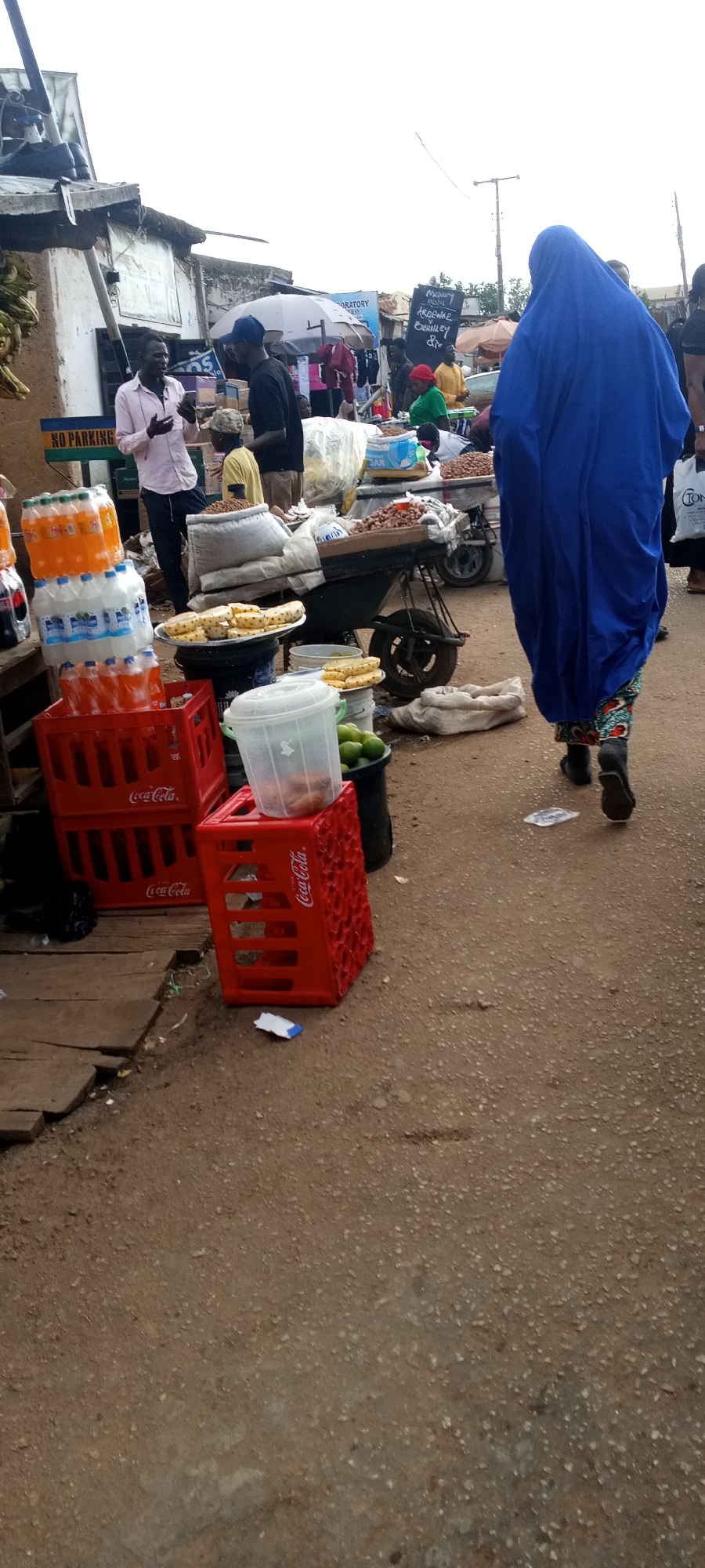 The image captures a bustling street scene at a market in an unknown location. A woman clad in a full blue gown and patterned leggings walks away from the camera, right of the frame. The market area is crowded, with various vendors and shoppers present. A man in a light pink shirt is engaged in a conversation, possibly on a mobile phone, standing near stacks of bottled beverages and Coca-Cola crates. Other individuals are visible, engaging in buying and selling activities around wheelbarrows laden with goods. A sign reading "NO PARKING" is attached to a building. Sign with letters "ORATORY", "ARORNGR" and "BERILLEY" is also visible. The ground is uneven and appears to be a mix of dirt and gravel.