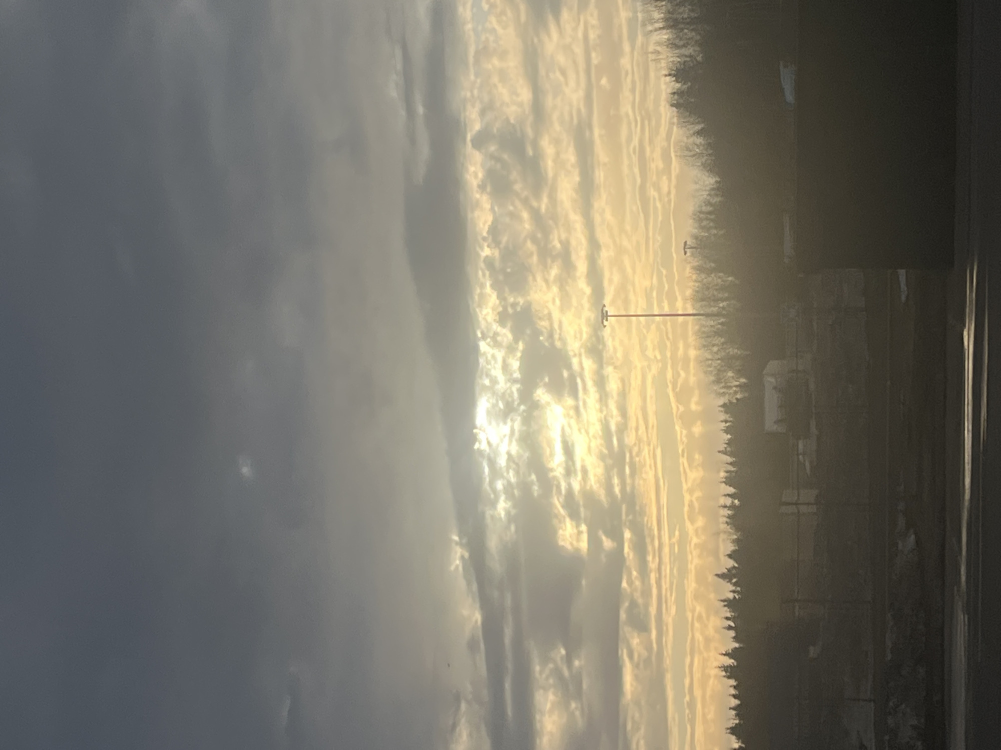 The image shows a landscape at sunrise or sunset, with a dramatic sky filled with textured clouds ranging from dark gray to bright golden hues. The sun is visible behind some clouds. The horizon features a line of trees in the middle ground, with an open field or area beyond, and a couple of light poles are seen. A dark structure is present on the right edge of the frame, obscuring part of the scene. The environment suggests a location outside a city area. The scene features neither people nor discernible interactions. The location, Lupanovo, Russia, is not identifiable from the image.