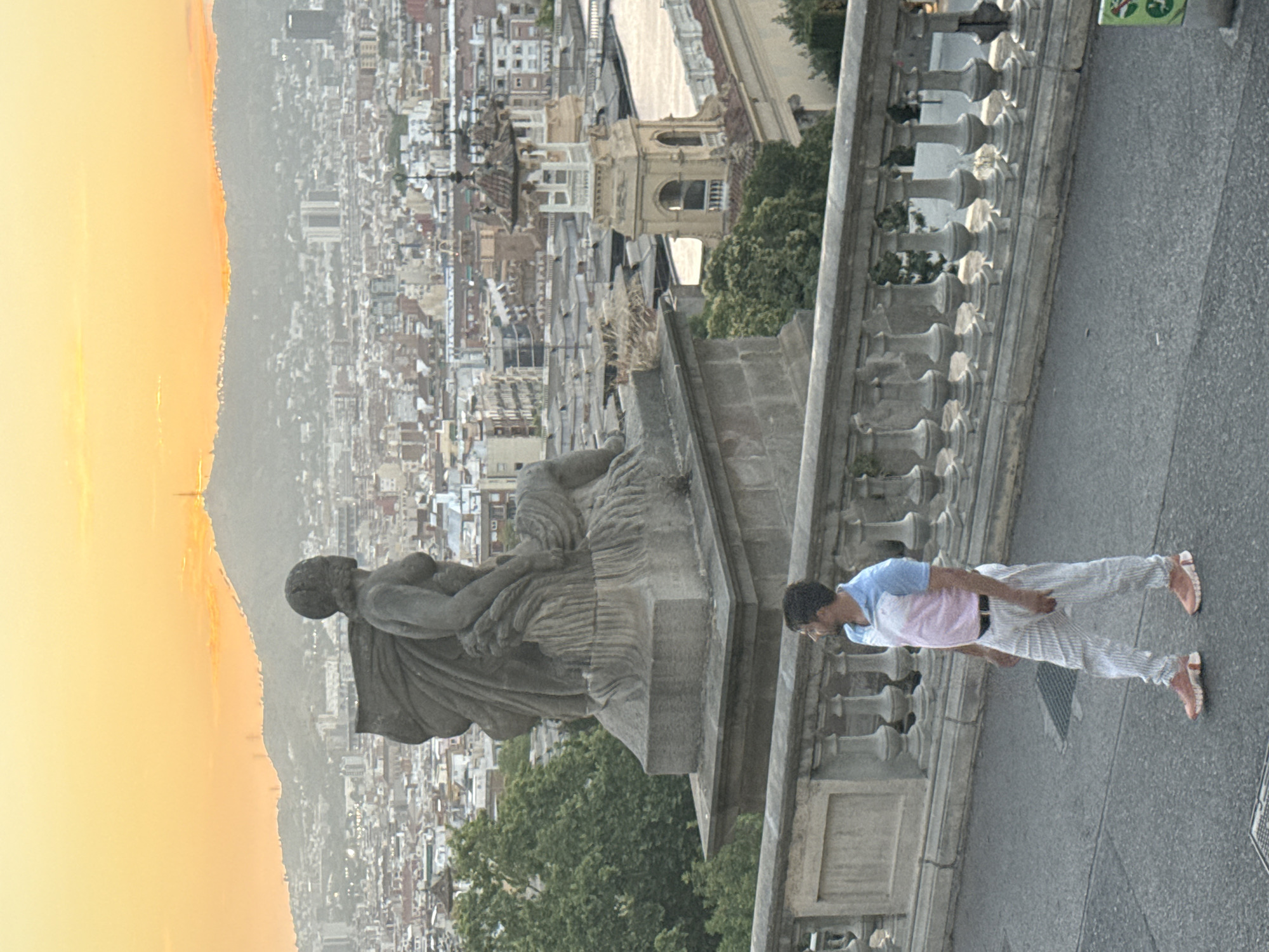 The image captures a panoramic view of Barcelona, Spain, at sunset. In the foreground, a man stands on a paved walkway, looking towards the city. He is wearing a pink and blue polo shirt, white striped pants, and pink sneakers. Behind him, a stone balustrade with ornate columns runs along the edge of the walkway. To the left of the man, a large stone statue of a seated figure, possibly a mythological goddess, is prominent. The statue is weathered and appears to be part of a monument.
The midground features the sprawling cityscape of Barcelona, characterized by a dense collection of buildings with terracotta roofs. Interspersed within the urban landscape are several notable architectural elements, including church spires and towers, one of which has a distinctive yellow facade and intricate detailing. Green trees are visible in patches throughout the city.
The background is dominated by a hazy, orange-hued sky, typical of a sunset. In the distance, a large, tree-covered hill rises, with a visible structure on its summit, likely a communication tower or landmark. The overall atmosphere is serene, with the warm light of the setting sun casting a soft glow over the scene.
