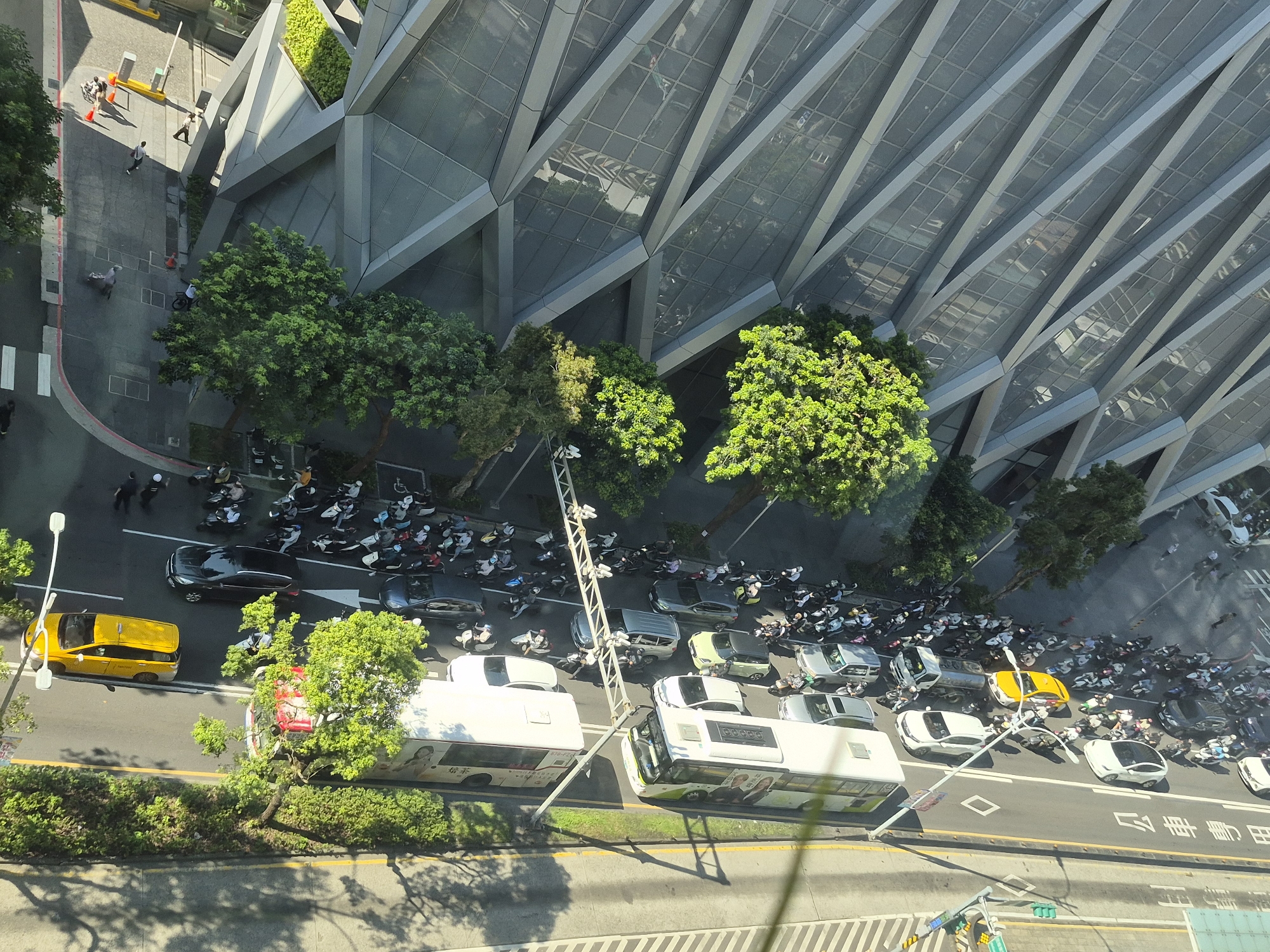 The image captures a street scene in Taipei, Taiwan, looking down from an elevated perspective. A large, modern building with a distinctive geometric facade dominates the upper portion of the frame. In the foreground, a street is filled with various vehicles, including a yellow taxi, several cars, and a large number of motorcycles and scooters parked along the side and in a lot. Two buses are also visible on the road. Pedestrians can be seen walking on the sidewalk and near the building entrance. The street is lined with trees, and shadows are cast across the road, indicating it is daytime. The scene appears to be a busy urban environment.