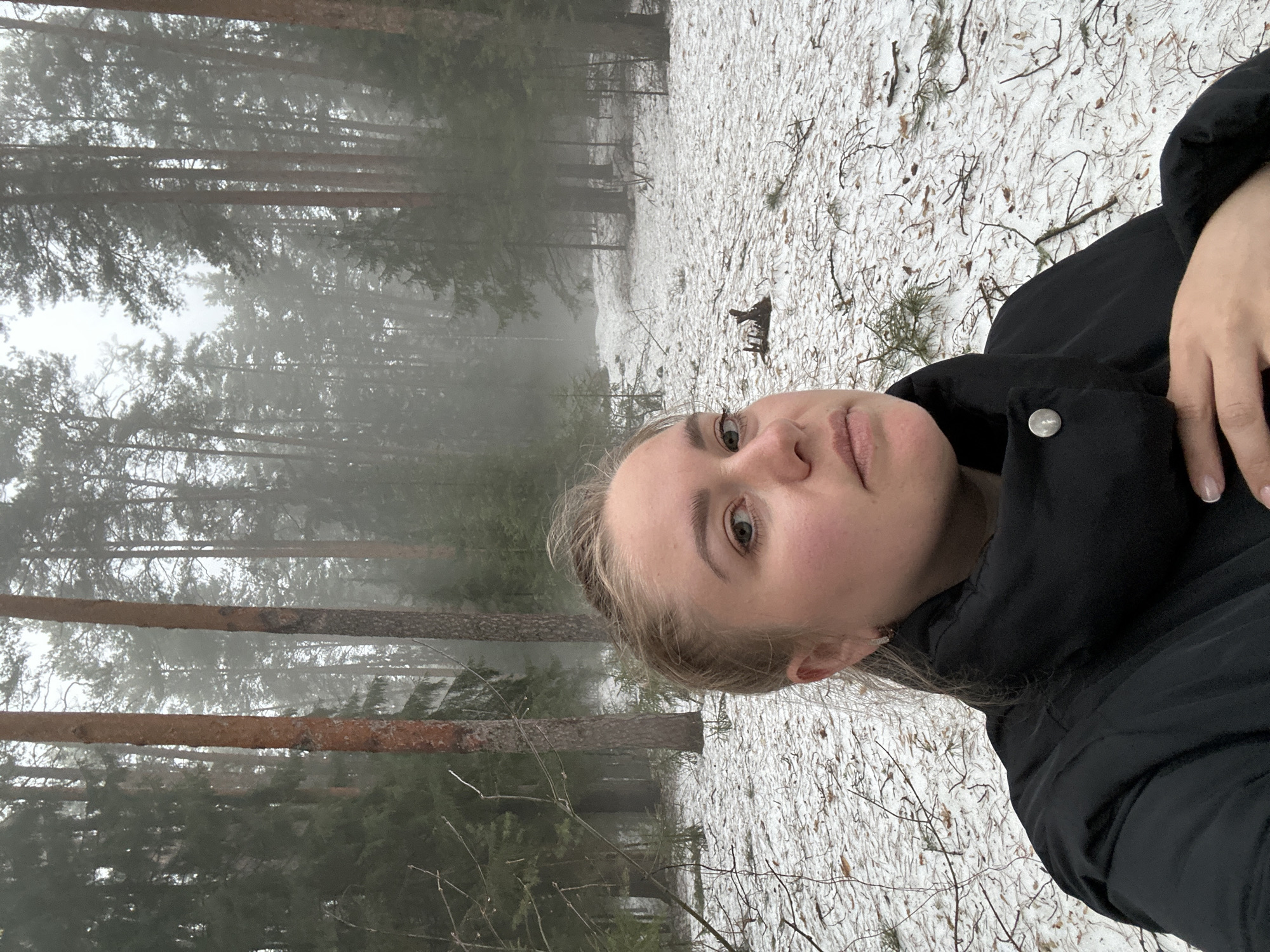 A selfie taken outdoors in a wooded area with sparse snow cover on the ground. Tall trees are visible in the background, obscured by fog. The subject is a woman with fair skin and blonde hair, wearing a black jacket. The location appears to be a forest, possibly in a park or outskirts of a city. There is no indication of Chaikovsky, Russia, beyond the visual cues of a cold climate and a forest environment. No specific actions or interactions are depicted, other than the subject taking the selfie.
