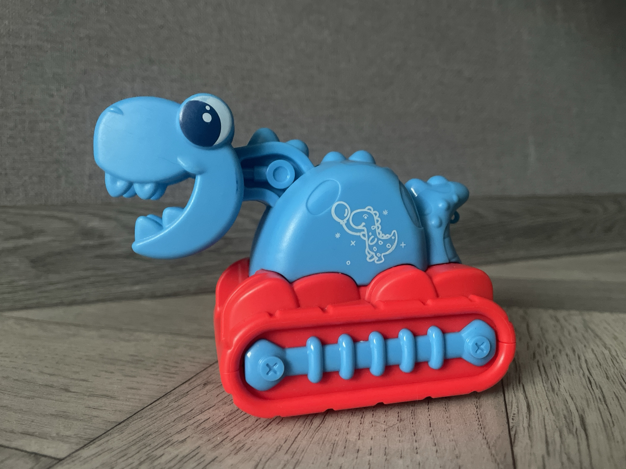 The image shows a plastic toy dinosaur bulldozer. The dinosaur head and body are light blue with a white dinosaur print on the side. The bulldozer tracks are red with blue rollers and screws. The toy sits on a wood-patterned surface against a gray wall. The location is Unknown, Unknown.
