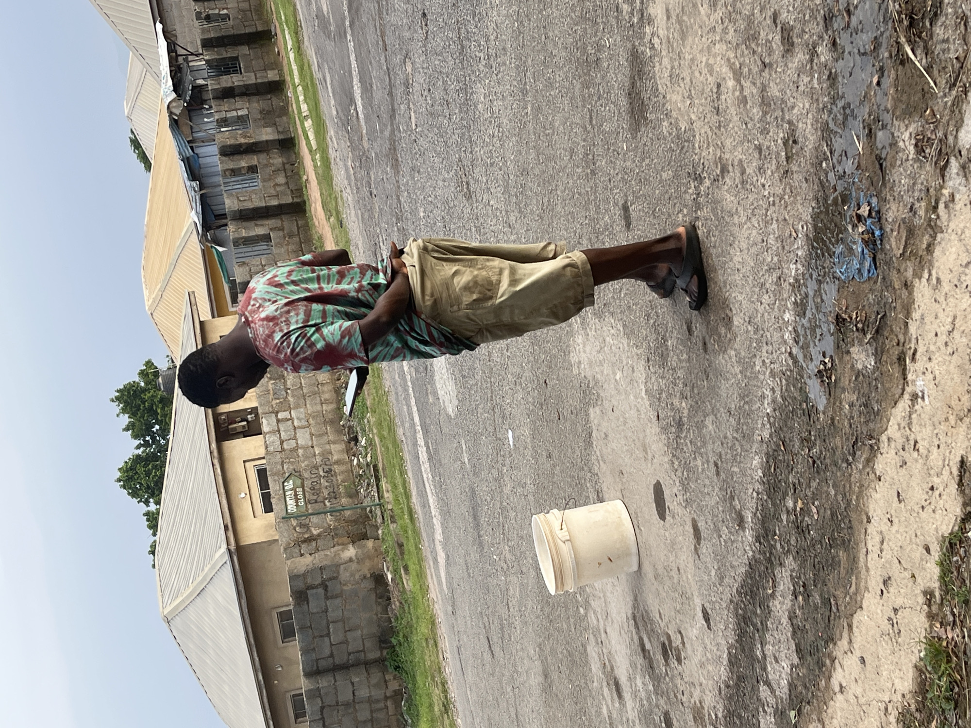 In Abuja, Nigeria, a young Black male stands on a paved street, facing away from the camera. He is wearing a tie-dye t-shirt in shades of green and red, and khaki shorts. He is looking down at a tablet computer held in his hands. To his left, a beige bucket stands on the ground. In the background, several buildings with corrugated metal roofs are visible, along with a brick wall. A street sign in the background points to "ODUNTAN O.G. CLOSE" and displays a phone number. The ground is a mix of asphalt and dirt, with some puddles and scattered debris. The sky is overcast.