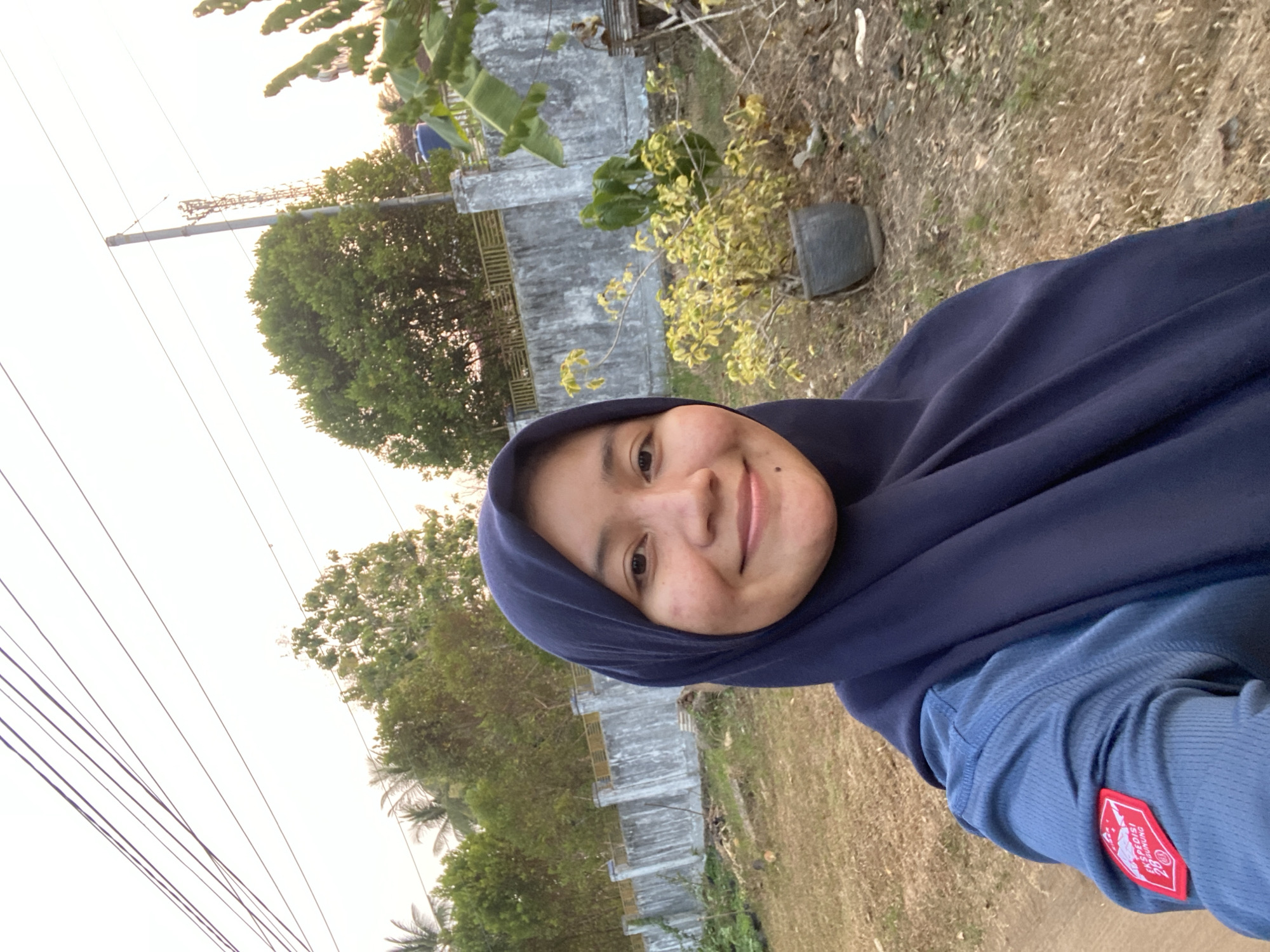 A young woman wearing a navy blue hijab and a blue long-sleeved shirt is taking a selfie outdoors. She has a slight smile and is looking towards the camera. Her shirt has a red patch on the left sleeve with white text and a mountain graphic. The background shows a dry, grassy area with scattered debris, a concrete wall with a yellow railing, and several trees. In the distance, a cell tower is visible. The sky is overcast with a warm, sunset glow. The setting appears to be a rural or semi-urban area in Banjarbaru, Indonesia, possibly near a residential or undeveloped area.