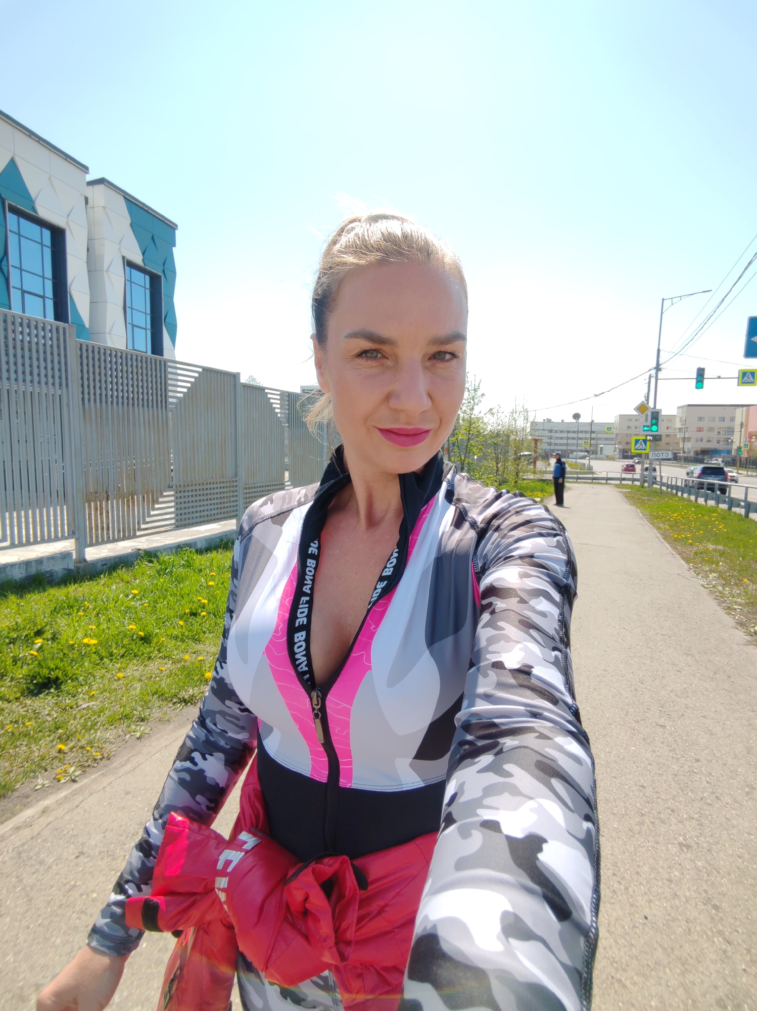 A woman, approximately 30-40 years old, with light hair pulled back, green eyes, and pink lipstick, captures a selfie on a sunny day in Petropavlovsk-Kamchatsky, Russia. She wears a long-sleeved athletic top featuring a grey, white, and black camouflage pattern with a prominent pink stripe and a black zippered front. A bright red puffer jacket is tied around her waist. The reversed text "BONA FIDE BONA" is visible on a black band of her top. She stands on a concrete sidewalk, bordered by a strip of grass with yellow dandelions to her left. Adjacent to the grass is a modern metal fence, beyond which stands a multi-story building with a white facade adorned with a blue-green geometric pattern and large, dark-framed windows. To the woman's right, a multi-lane street is active with vehicles, including cars and an SUV. Multiple traffic lights display green signals, and a pedestrian crossing sign with partially visible Cyrillic text ("ПОТО") is present. A lone pedestrian in a dark jacket is visible on the opposite side of the street. Utility poles with overhead wires and various urban buildings extend into the background beneath a clear blue sky.