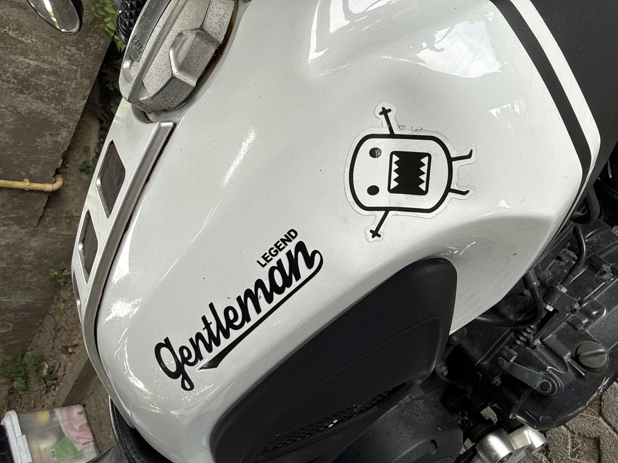 The image shows a close-up of a white motorcycle fuel tank. A black sticker with the word "Gentleman Legend" in a cursive font is prominently displayed on the tank. To the right of this sticker is a smaller black sticker of a cartoon character with wide eyes and a large, jagged mouth, resembling Domo-kun. The motorcycle is parked outdoors, with a concrete wall and some vegetation visible in the background. The scene appears to be in an urban or semi-urban environment. No people are visible in the image.