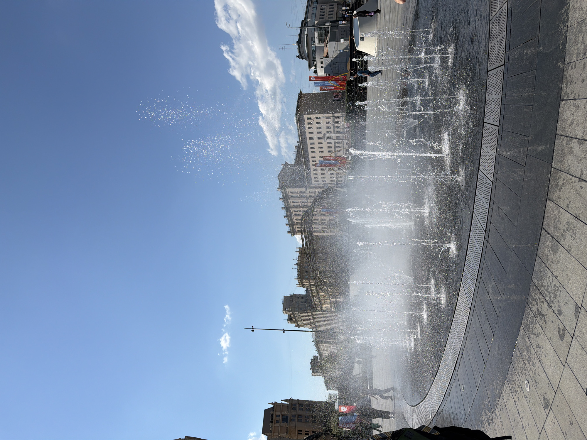 The image shows a fountain in a public square in Moscow, Russia. The fountain is circular, with jets of water spraying upwards. The ground around the fountain is paved with gray tiles. Buildings are visible in the background, including a building with a sign that reads "НОМЕРА МН". There are people walking near and through the fountain. The sky is blue with some white clouds.