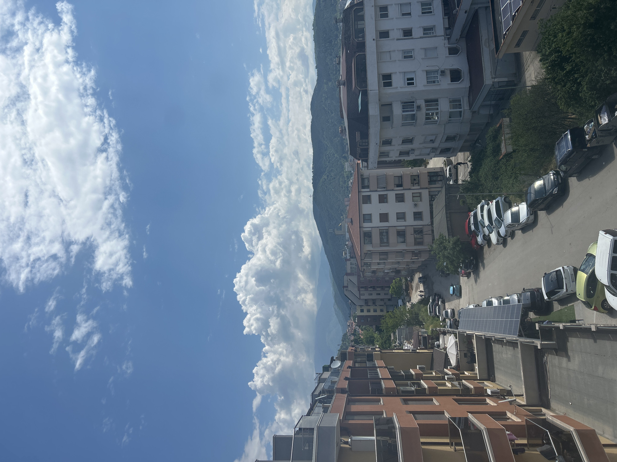 The image is a vertical outdoor shot taken from an elevated position, possibly a balcony or high-rise building, in what appears to be the city of Blagoevgrad, Bulgaria.
The view encompasses a street with several parked cars on the right side. The architecture consists primarily of residential buildings of varying heights, most of which are medium to high-rise. The buildings have a mix of architectural styles, with some showing signs of age.
In the background, a mountain range is visible, partially obscured by the buildings. The left side of the image is dominated by a blue sky with puffy white clouds.
No people are distinctly visible in the image. The environment is urban, with elements of nature (mountains, sky) incorporated into the scene.