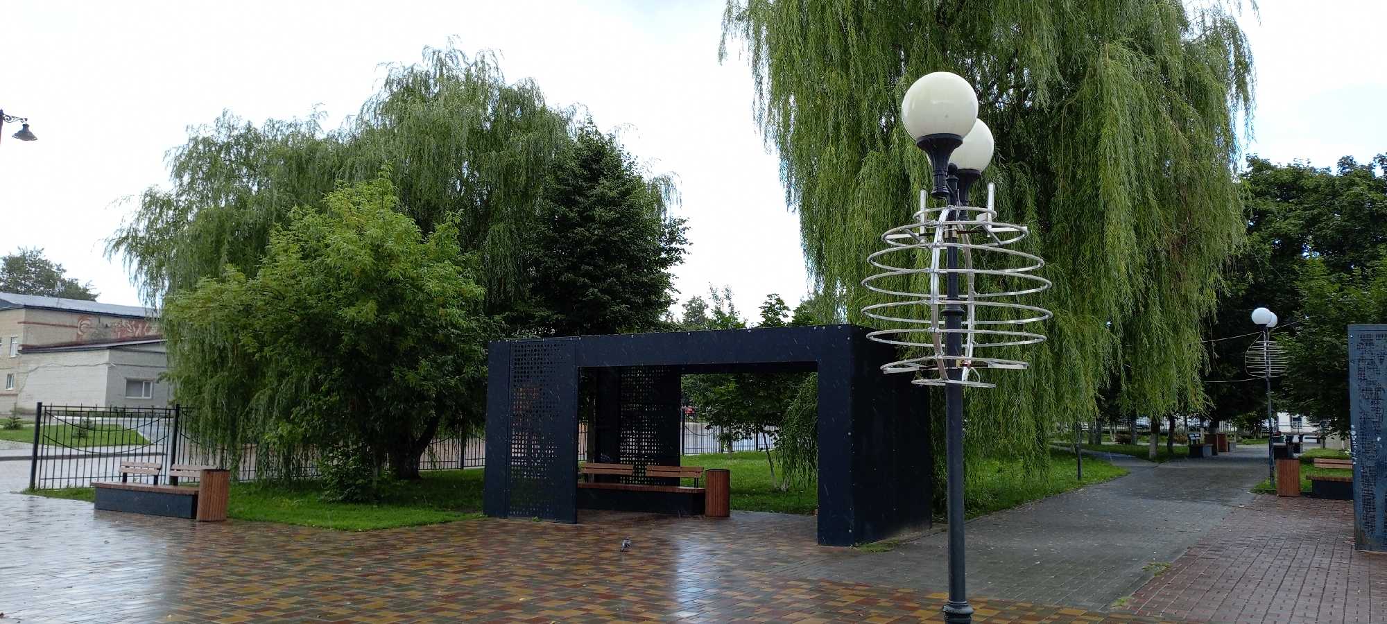 The image shows a public park area in the city of Kirsanov, Russia. In the foreground, a wet, paved walkway with a pattern of colored rectangular bricks leads into the scene. To the left, a bench with a wooden seat and a dark base is situated next to a wooden trash receptacle. Behind this, a black metal fence encloses a grassy area with a smaller tree. Further back, a two-story building with a light-colored facade and some artwork on the upper section is visible.
Dominating the mid-ground is a dark, modern, open-sided pavilion with a slatted roof and walls featuring decorative circular perforations. Inside the pavilion, another bench with a wooden seat is visible.
To the right of the pavilion stands a tall, dark lamppost with two white spherical lamps. This lamppost is adorned with a distinctive silver, spiraling decorative element.
The background is filled with lush green trees, primarily large weeping willows with their characteristic drooping branches, and other deciduous trees. The sky is overcast, suggesting a cloudy day. A single pigeon is visible on the wet pavement in front of the pavilion. The scene appears to be a tranquil, well-maintained public space.