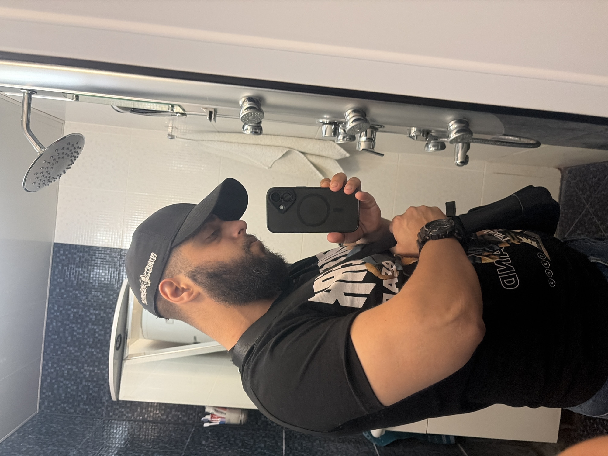 A selfie captured in a bathroom setting. The individual taking the photo is a man with a beard, wearing a black cap and a black t-shirt. He's holding a smartphone in his right hand, presumably to take the picture. The bathroom contains a showerhead, faucets, a toilet, and tiled walls. The city and location are unknown.