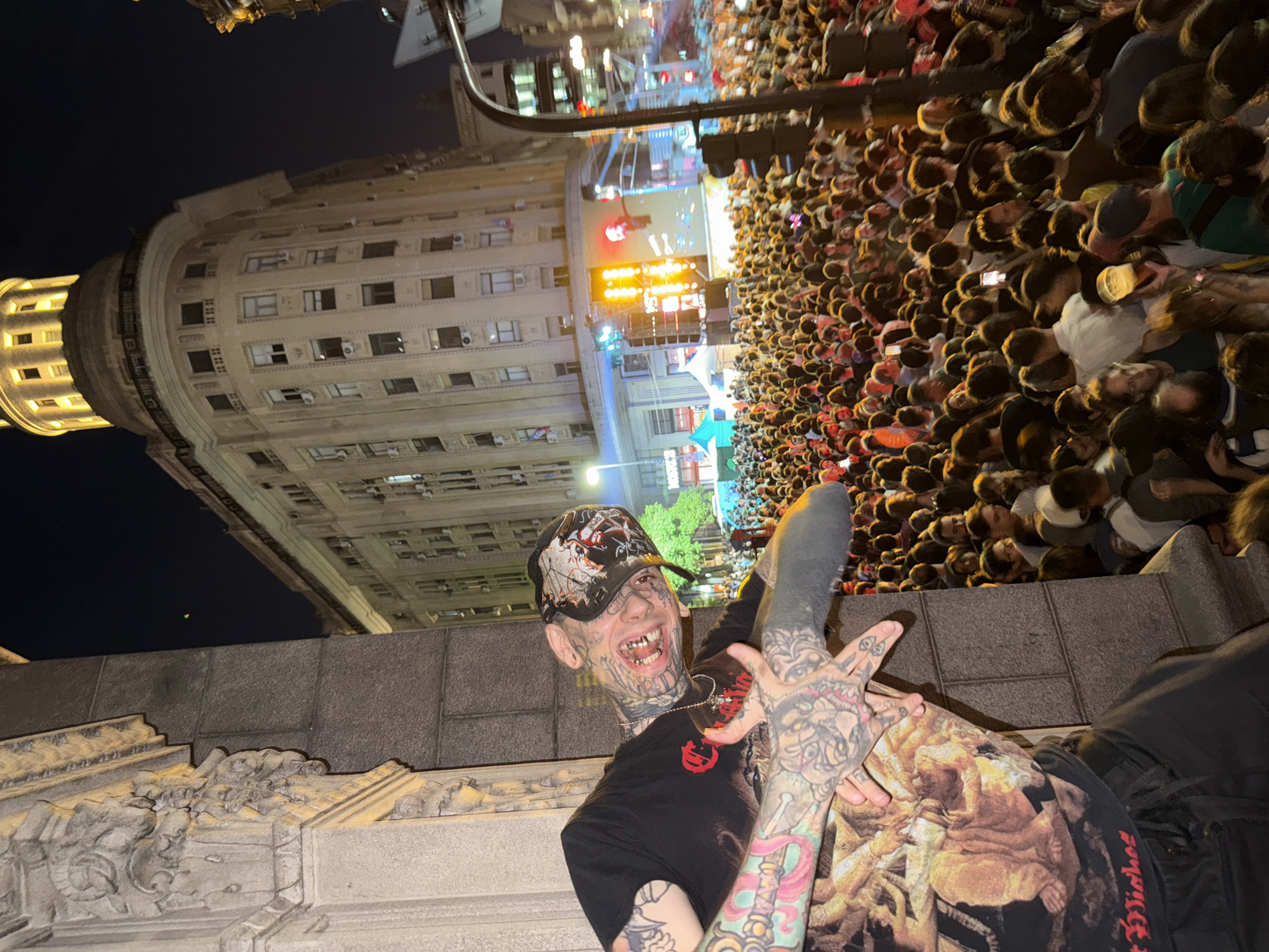 The image depicts a man with tattoos and a hat, posing in front of a large crowd gathered in a city street at night.
The man is wearing a black T-shirt, tattoos covering his face and arms. His expression includes a visible smile. He is making a hand gesture towards the viewer.
Behind the man is a dense crowd of people. The crowd is focused towards a stage with a large screen displaying lights and figures. The environment suggests a concert or a large public event.
In the background are illuminated buildings, typical of urban architecture.
Based on the image elements and the presence of a large crowd, it is highly likely that this scene is in the city of Buenos Aires, Argentina, during a public event, potentially near a landmark building.