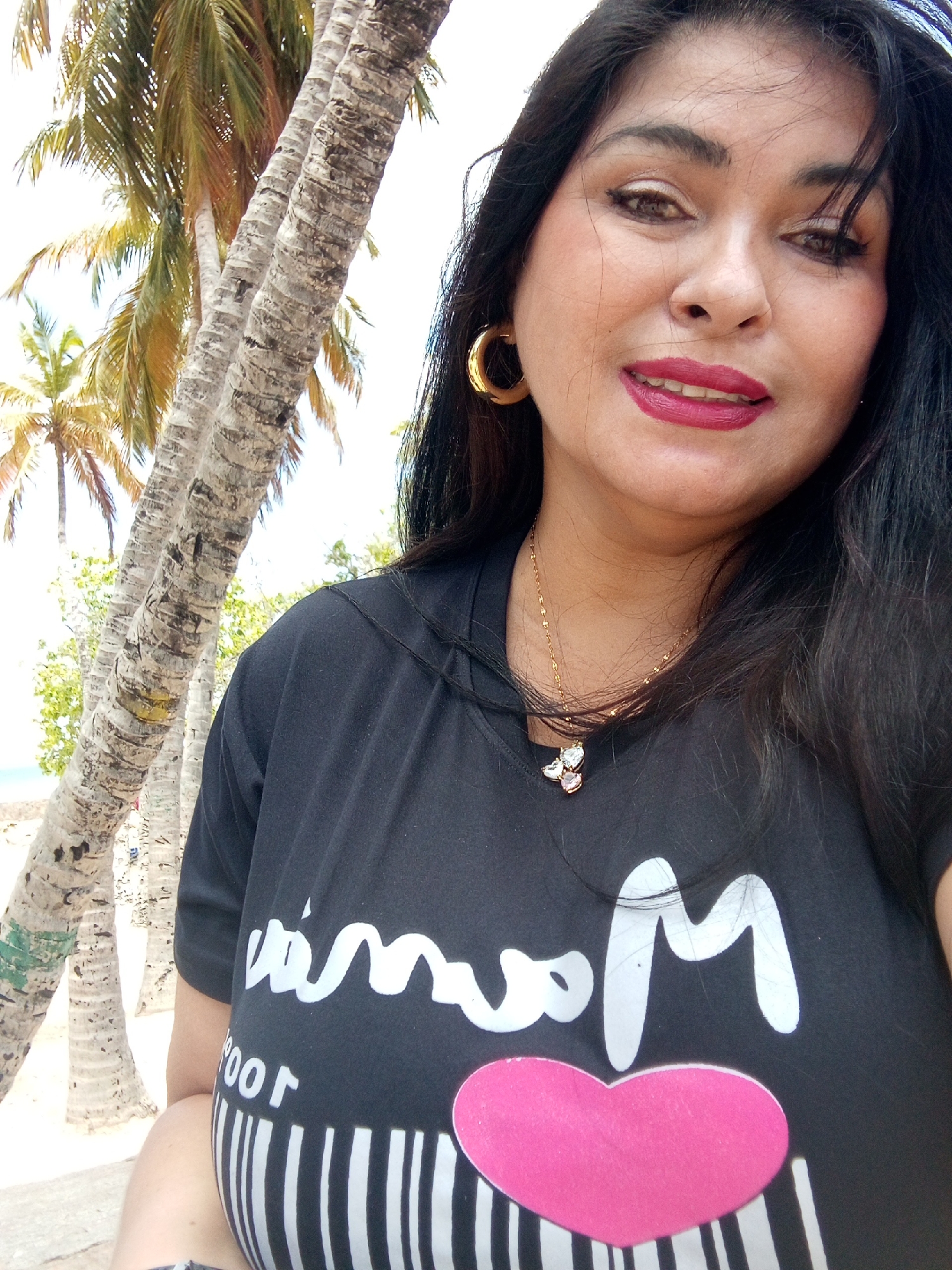 A person is taking a selfie outdoors in an unknown location in the city of Unknown, Unknown. Palm trees are visible in the background. The person has dark hair, is wearing gold hoop earrings and a gold necklace with a pendant. The person is wearing a black t-shirt with white lettering and a pink heart design.