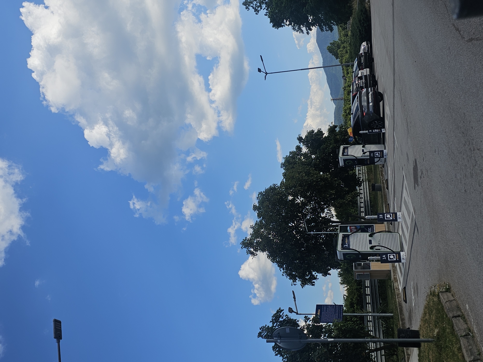 The image shows an outdoor scene, likely a parking lot for electric vehicles, with a clear blue sky and scattered white clouds. In the foreground, there are several electric vehicle charging stations, some labeled with "1A" and "1B" and bearing a green charging symbol. The stations appear to be modern and white with green accents.
Behind the charging stations, a row of parked cars is visible, suggesting they are either charging or waiting to charge. The cars are dark in color, and some have license plates visible, though they are not legible enough to read.
In the background, lush green trees form a dense line, and beyond them, rolling hills or mountains are visible, indicating a scenic or rural location. The time of day appears to be daytime, given the bright sunlight and clear sky. The weather seems pleasant, with a bright sky and sunshine.
There is a lamppost visible in the sky on the left side of the image. Another lamppost with multiple lights is also present in the upper right portion of the image. A blue sign with white text is partially visible on the right side, possibly indicating information or instructions related to the charging station or parking area.
The scene depicts a functional setting for electric vehicle charging, set against a backdrop of natural beauty. No people are visible in the image. The overall mood is calm and orderly.