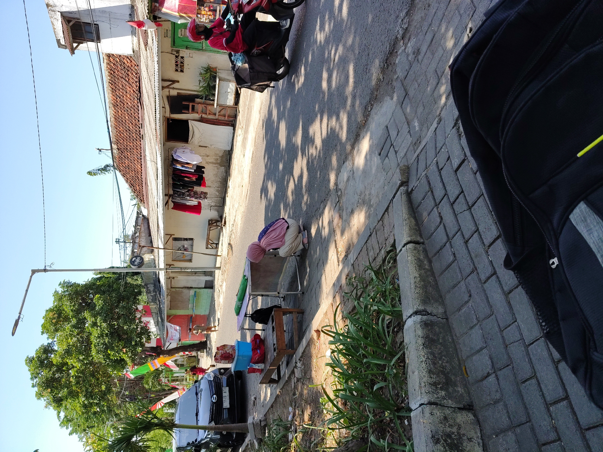 The image captures a street scene in Surabaya, Indonesia, on a bright, sunny day. In the foreground, a black backpack is partially visible, suggesting a personal perspective. A paved walkway lines the street, with a curb separating it from a patch of greenery. Beyond the walkway, a dark-colored car is parked, and further down the street, a woman wearing a pink hijab and beige pants is squatting next to a folding table. She appears to be interacting with the table's contents, which include a blue plastic container and bags.
A wooden bench with items on it sits beside the folding table. A second, smaller folding table is covered with a green tarp. The street itself is asphalt, with dappled sunlight casting shadows from overhead trees. A person on a red motorcycle with a black cargo box is riding away from the viewer down the street.
The background reveals a row of modest buildings with tiled roofs. Laundry is hanging on a line in front of one of the buildings, and Indonesian flags are displayed, indicating a possible celebration or national holiday. Two young boys are visible further down the street, one walking and another running. A tall street lamp stands prominently on the left side of the image. The overall impression is of a typical urban environment in Surabaya, bustling with everyday activity.