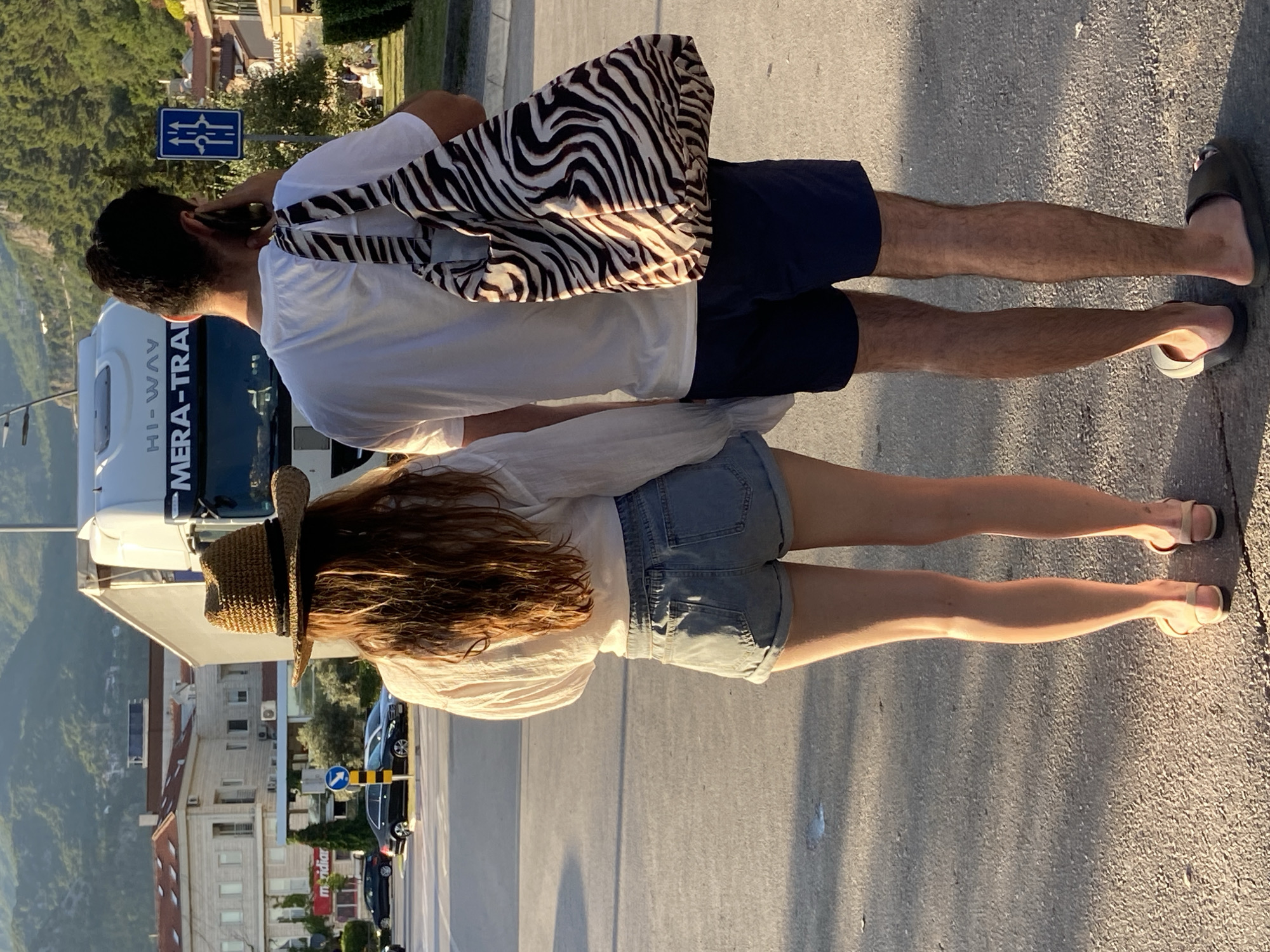 A full-length, back-view shot captures two individuals, a woman and a man, standing on an asphalt road in Budva, Montenegro, bathed in the warm light of what appears to be late afternoon. The woman, positioned on the left, wears a wide-brimmed straw hat, a loose white long-sleeved shirt, and light denim shorts. Her long, auburn hair cascades down her back, and she is wearing flat, beige sandals. The man, to her right, is dressed in a plain white t-shirt and dark blue shorts. He has a large tote bag with a black and white zebra print slung over his right shoulder. His left hand is raised, holding a dark-colored mobile phone to his ear, suggesting he is engaged in a conversation.
Behind them, a large white truck with "HI-WAY MERA-TRA" visible on its side dominates the background. The truck is on a road, likely a main thoroughfare, with other vehicles, including cars, visible in the distance. To the right of the truck, a blue traffic sign with white arrows indicates lane directions. Buildings, characterized by light-colored facades and red-tiled roofs, line the street, and lush green trees and hills form the backdrop under a clear sky. The ground beneath the individuals is a textured asphalt surface with visible cracks and patches of lighter colored material, indicating wear and tear. The setting suggests a street scene in a Mediterranean town, with the architectural style and mountainous terrain characteristic of Budva.