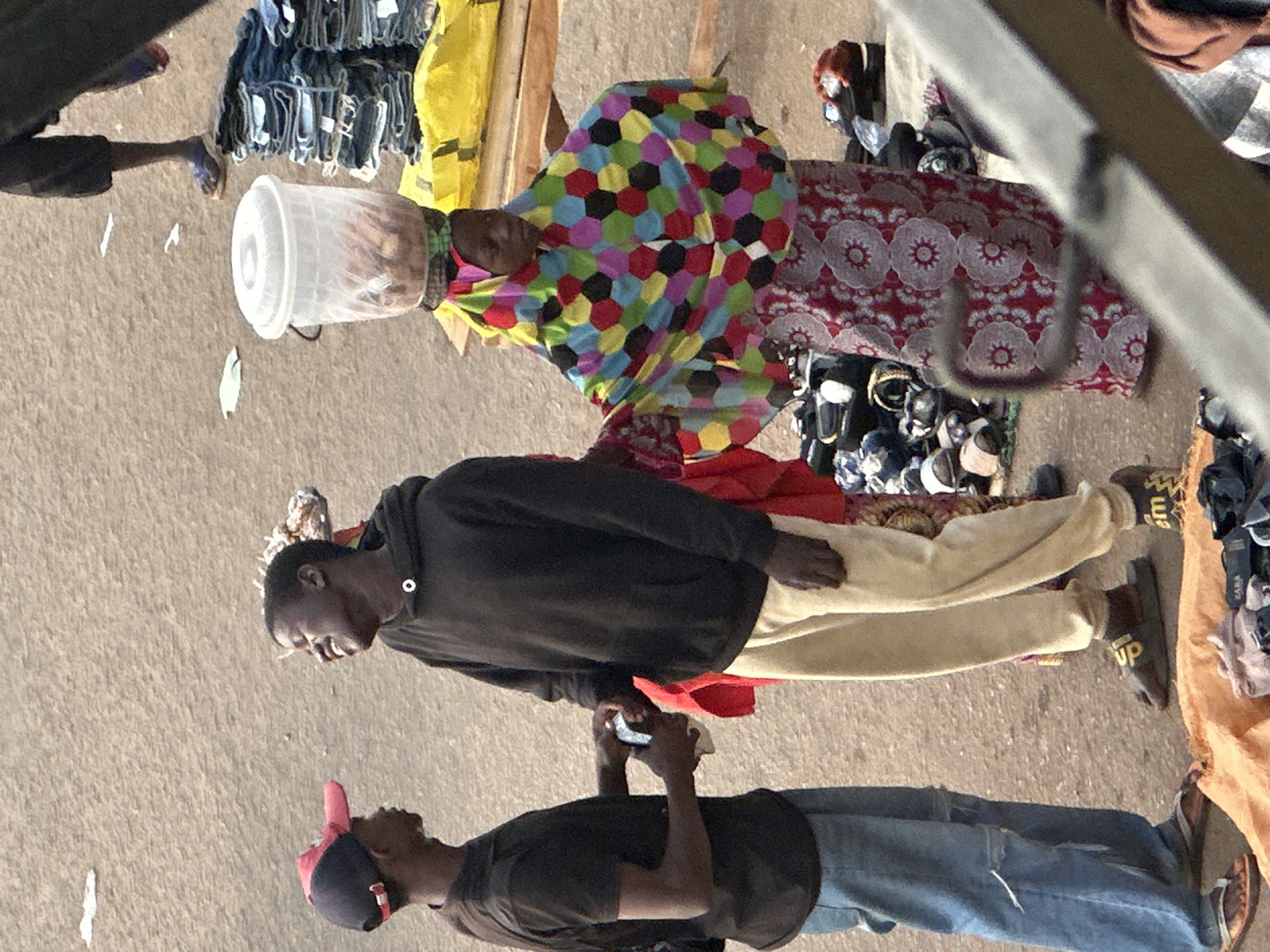 The image depicts an outdoor scene, likely a market, given the presence of various goods and individuals. The subjects are three people, two men and one woman, all appearing to be of African descent. The woman is wearing a brightly colored, hexagonal-patterned headscarf. One man is wearing a dark hoodie and light-colored pants, and the other is in a dark t-shirt and blue jeans.
The scene appears to be set during the day, with natural lighting. The ground is a sandy or dusty surface, consistent with an outdoor market setting. Visible objects include a white plastic bucket, stacks of clothing, and various footwear. One man is holding a white object, possibly a tissue or a small container.
The individuals are positioned in a way that suggests they are interacting or engaged in activities typical of a market. The man in the hoodie is looking down, while the man in the t-shirt is looking up and holding his hand up. The woman's gaze is directed downwards. The overall impression is one of everyday life and commerce.
There is no discernible text visible in the image. The mood is neutral, with no strong emotional cues evident from the subjects' expressions or postures. The location context is Bukuru, Nigeria, which aligns with the appearance of the people and the setting.