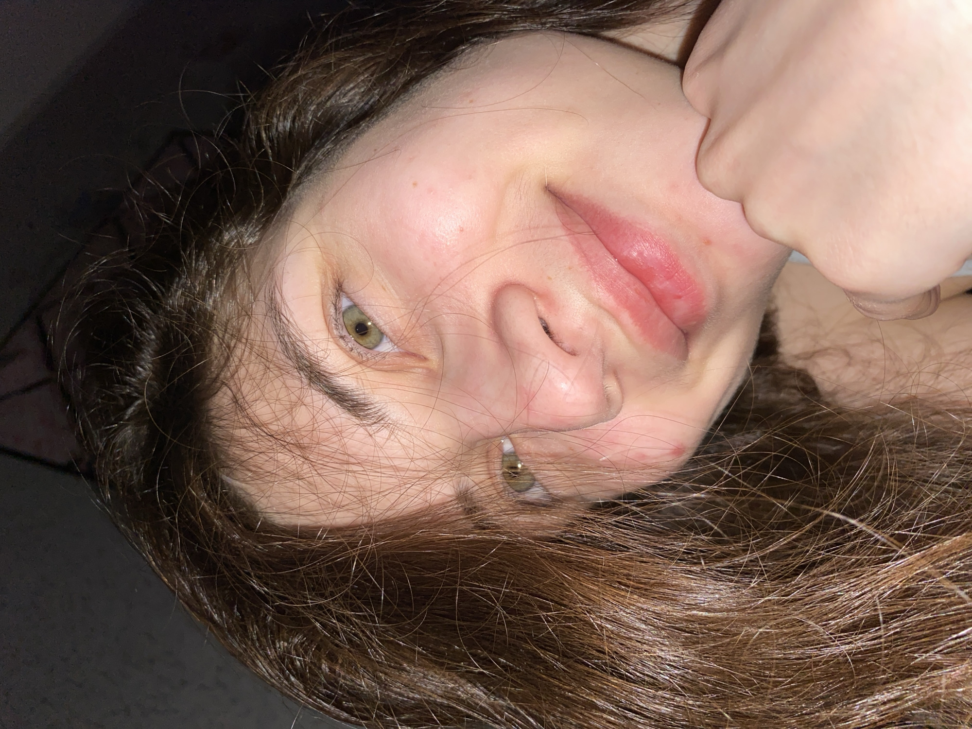 A rotated, close-up portrait features a person, likely female, with long, dark brown, wavy hair framing her face. She looks directly at the viewer with greenish-hazel eyes, her head tilted, and a slight, playful smile on her lips. One hand is positioned near her chin or jawline. Her skin appears fair, with some minor redness on her cheeks and around her mouth. The background is dark and out of focus, suggesting an indoor setting with dim lighting. The activity captured is a direct gaze at the camera, conveying a casual and perhaps coy emotion. No text is visible in the image. The provided location context for this media is Votkinsk, Russia.