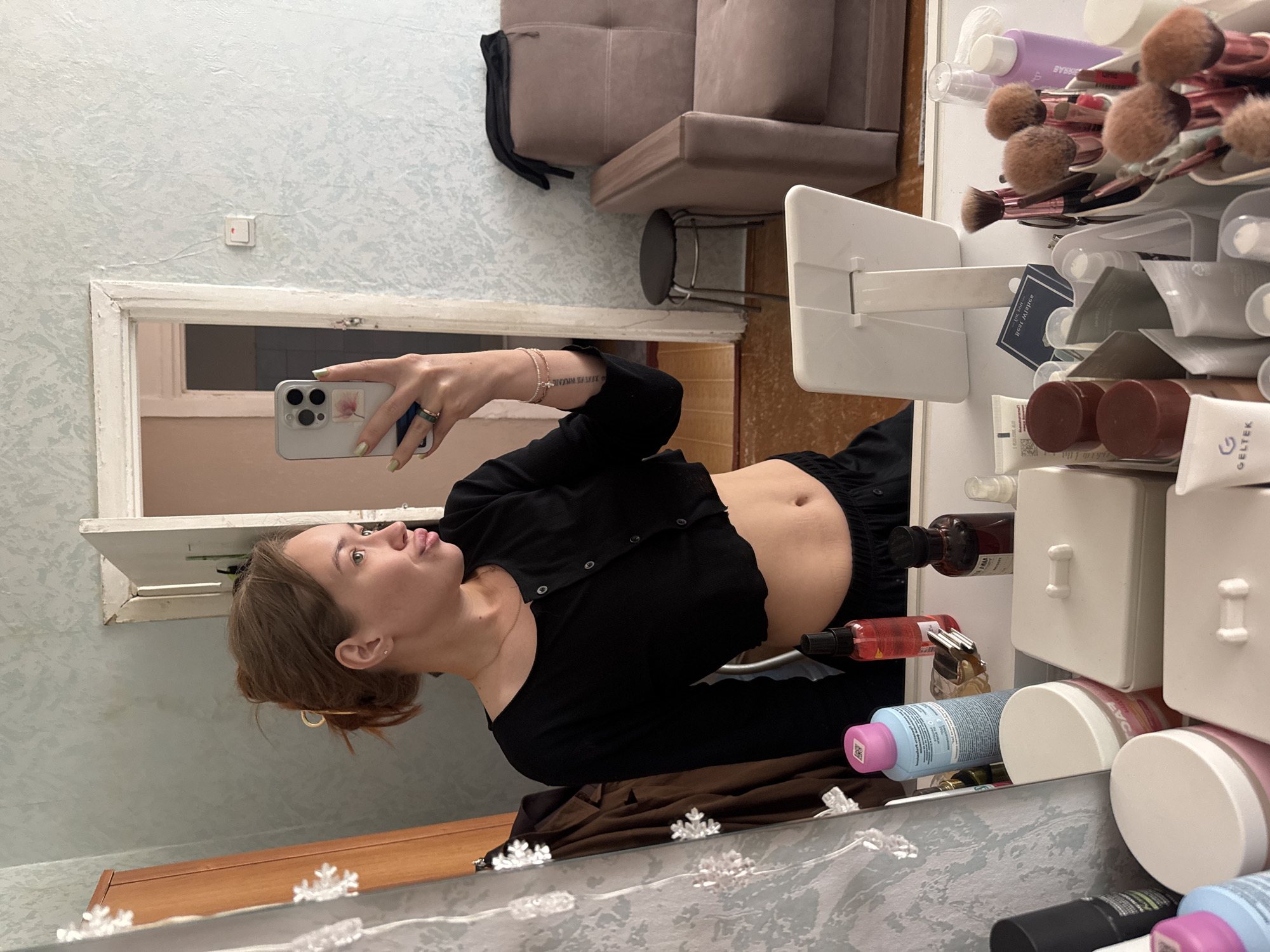 A young woman is taking a selfie in a mirror in a room. She is wearing a black cropped long-sleeved shirt and black pants. Her hair is pulled up in a bun. She is standing in front of a vanity table with various cosmetic items on it. The room appears to be a bedroom or dressing area, with a closet and a sofa visible in the background. The image does not contain enough information to determine the exact location within the city of Chaikovsky, Russia.