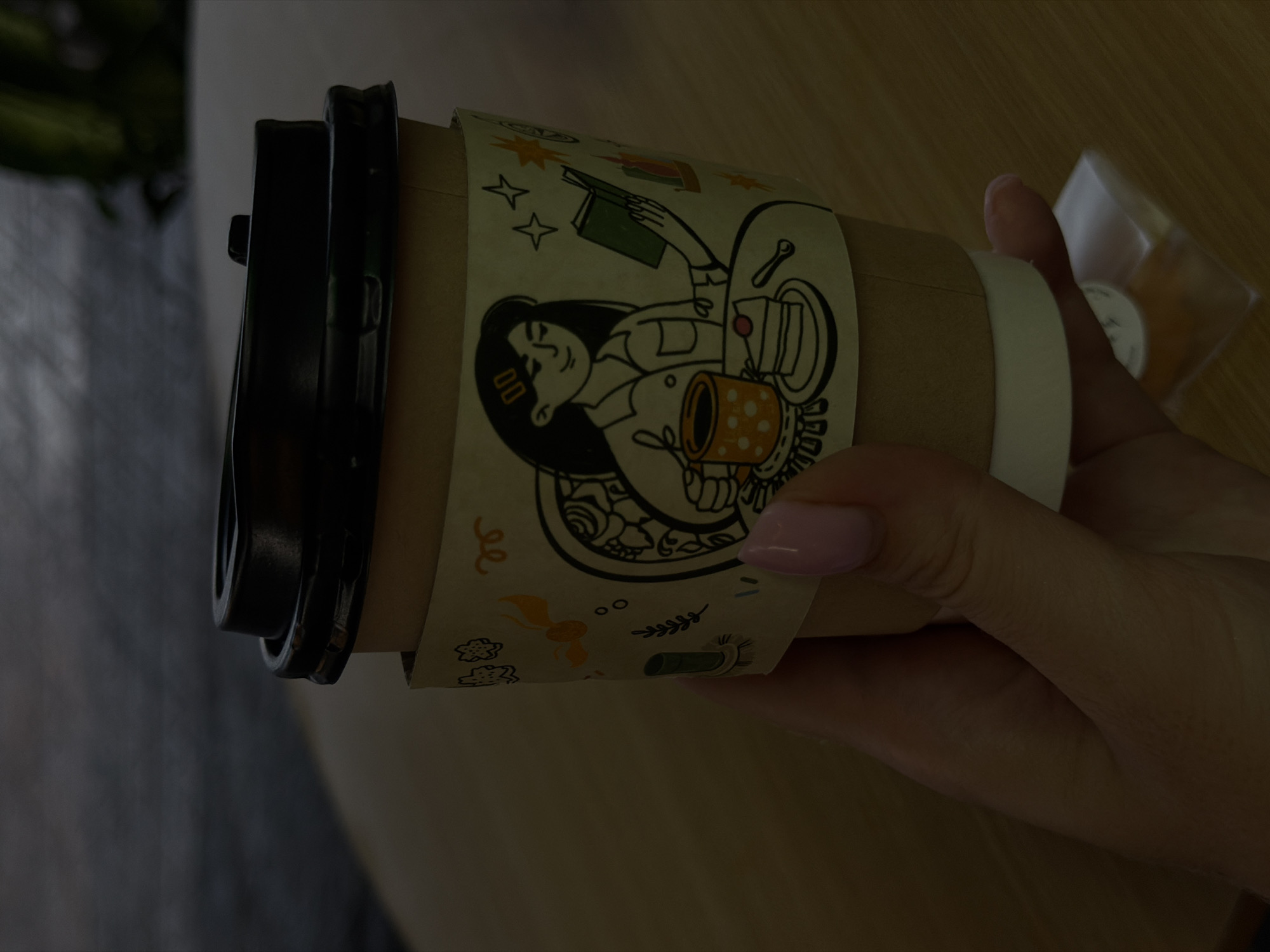 The image shows a hand holding a brown paper coffee cup with a black lid and a light-colored decorative band. The band features a cartoon illustration of a person with a cup of coffee and food. A clear plastic package with a small, circular object inside is visible in the background. The setting is indoors and the lighting is soft, with the background appearing to be a table. It's difficult to confirm the location within Bac Nha Trang Ward, Vietnam, as there are no specific landmarks or other location-defining elements visible in the image.