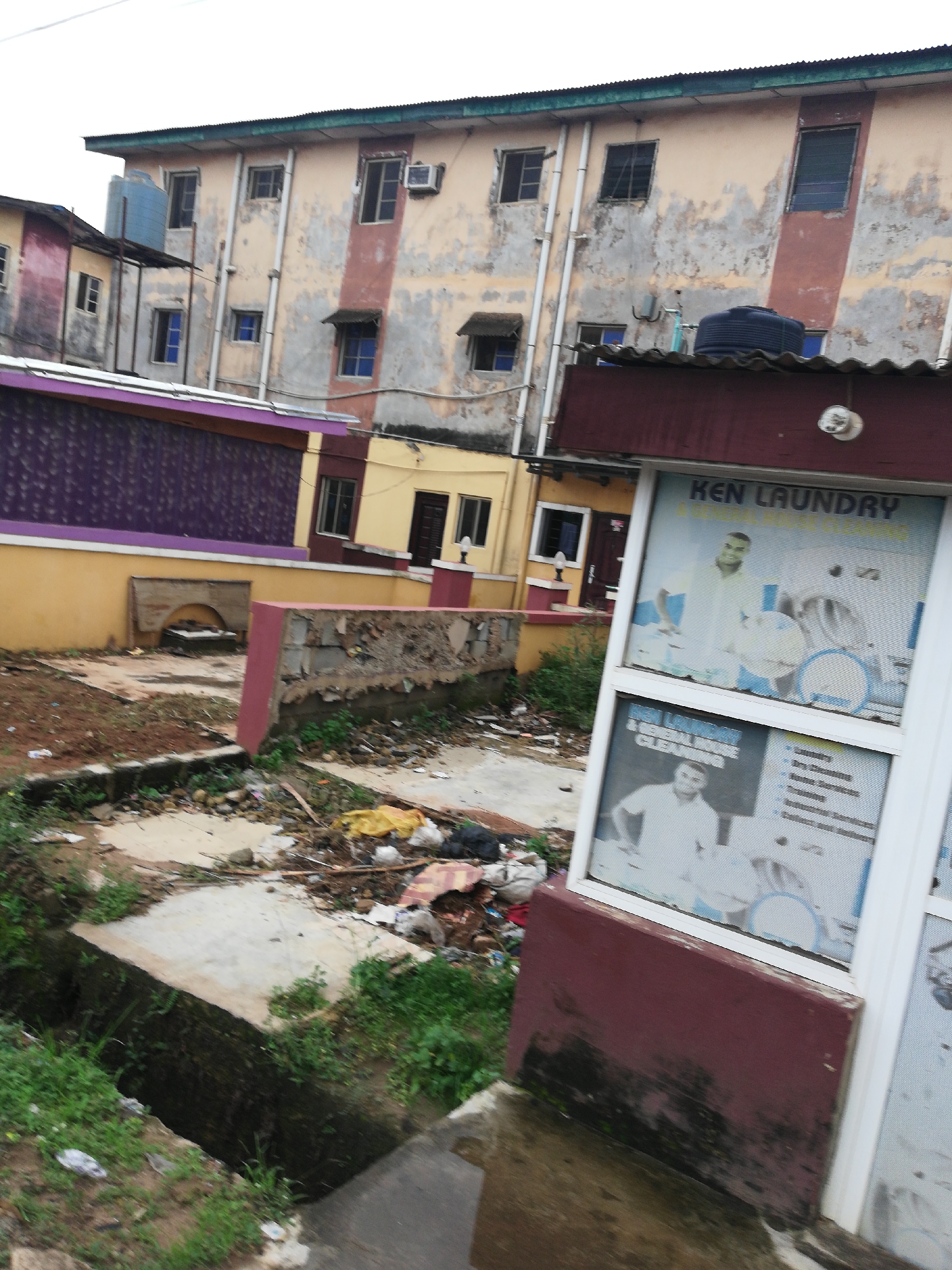 The image depicts an outdoor scene in what appears to be a neglected urban area. In the foreground, there is a drainage ditch with overgrown grass and visible trash. Behind the ditch, a plot of land shows signs of disrepair with broken concrete and more scattered trash. The middle ground features buildings in varying states of upkeep, including a two-story structure with purple and yellow accents and a taller, multi-story building with peeling paint and a generally run-down appearance. In the foreground, partially obscuring the view, is a window or sign for "KEN LAUNDRY," displaying promotional imagery of a man and a washing machine. The overall impression is one of urban decay and neglect. The location is in the city of Unknown, Unknown.