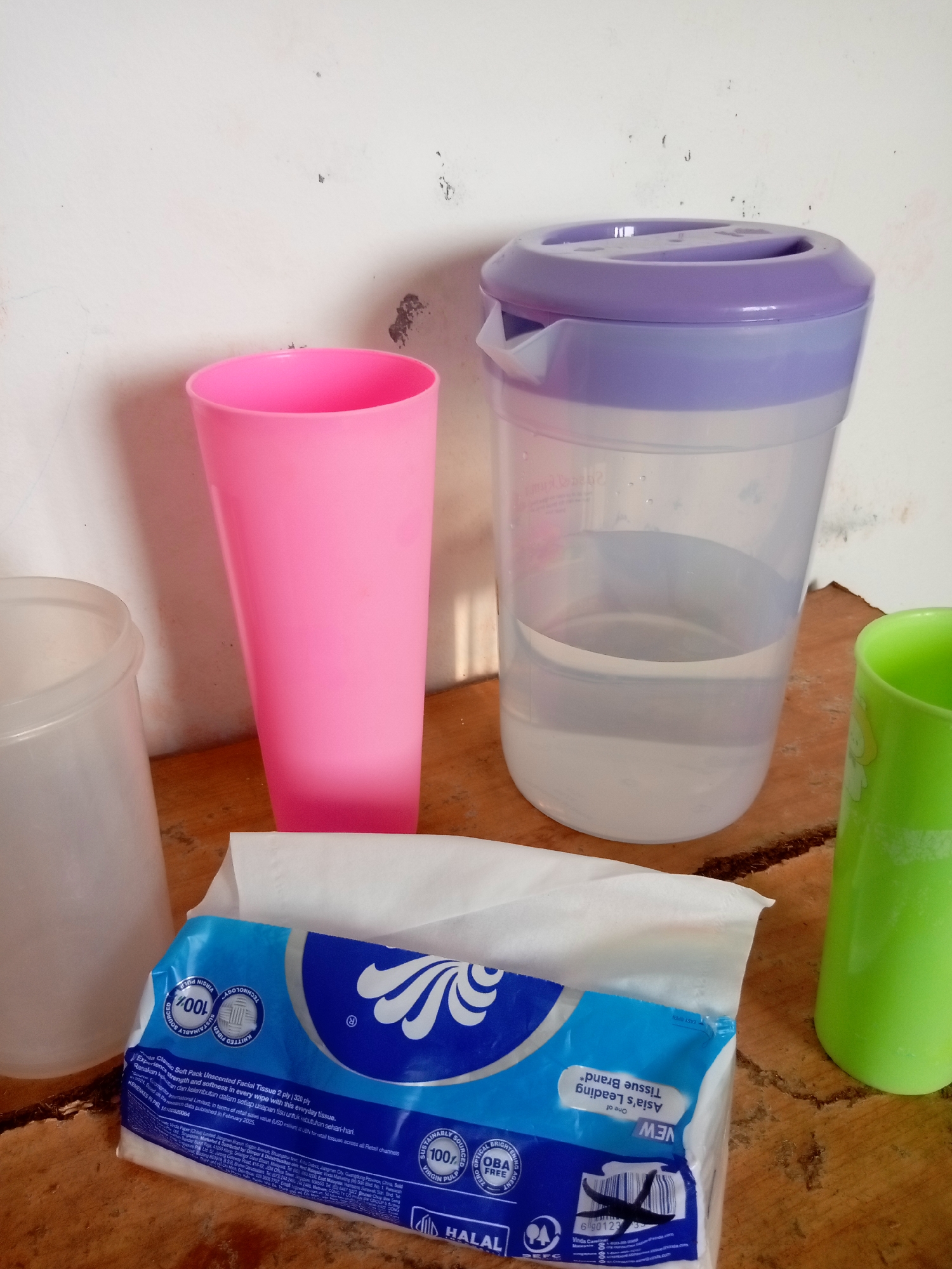 The image shows a still life of everyday objects placed on a wooden surface against a textured white wall. The scene is indoors, likely in a home or office setting in Batam, Indonesia, given the items present and typical Indonesian household items.
In the foreground, a pack of facial tissues with blue and white packaging is prominently displayed. The packaging features text and logos indicating "100% Virgin Pulp", "Fibre", and "Halal" certification, suggesting it is a consumer product.
Behind the tissues, to the left, is a tall, bright pink plastic cup. To its right is a large, clear plastic pitcher with a purple lid, containing a significant amount of clear liquid, likely water. Further to the right, a bright green plastic cup is visible. To the far left, partially obscured, is another white plastic container, possibly a smaller cup or jar.
The lighting in the image suggests it was taken during the daytime, with soft shadows cast by the objects. The background wall shows signs of wear, with some discoloration and texture, indicating a non-pristine environment. The wooden surface beneath the objects appears to be a table or countertop, with visible grain and some imperfections. No people are present in the image. The arrangement of the objects suggests a casual placement rather than a deliberate arrangement for photography.