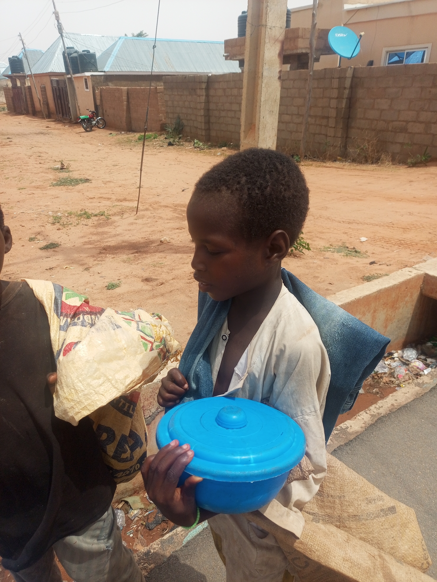 A young Black child in Tempure, Nigeria stands outdoors wearing a light-colored shirt and a dark denim jacket over their shoulder. They are carrying a blue plastic container with a lid in their hands. Another person, partially visible, stands to the left carrying a patterned sack. The setting appears to be a street with buildings and a motorcycle visible in the background. There is trash visible alongside the curb. The ground is mostly bare dirt with some sparse vegetation. A DStv satellite dish is mounted on a building in the background.
