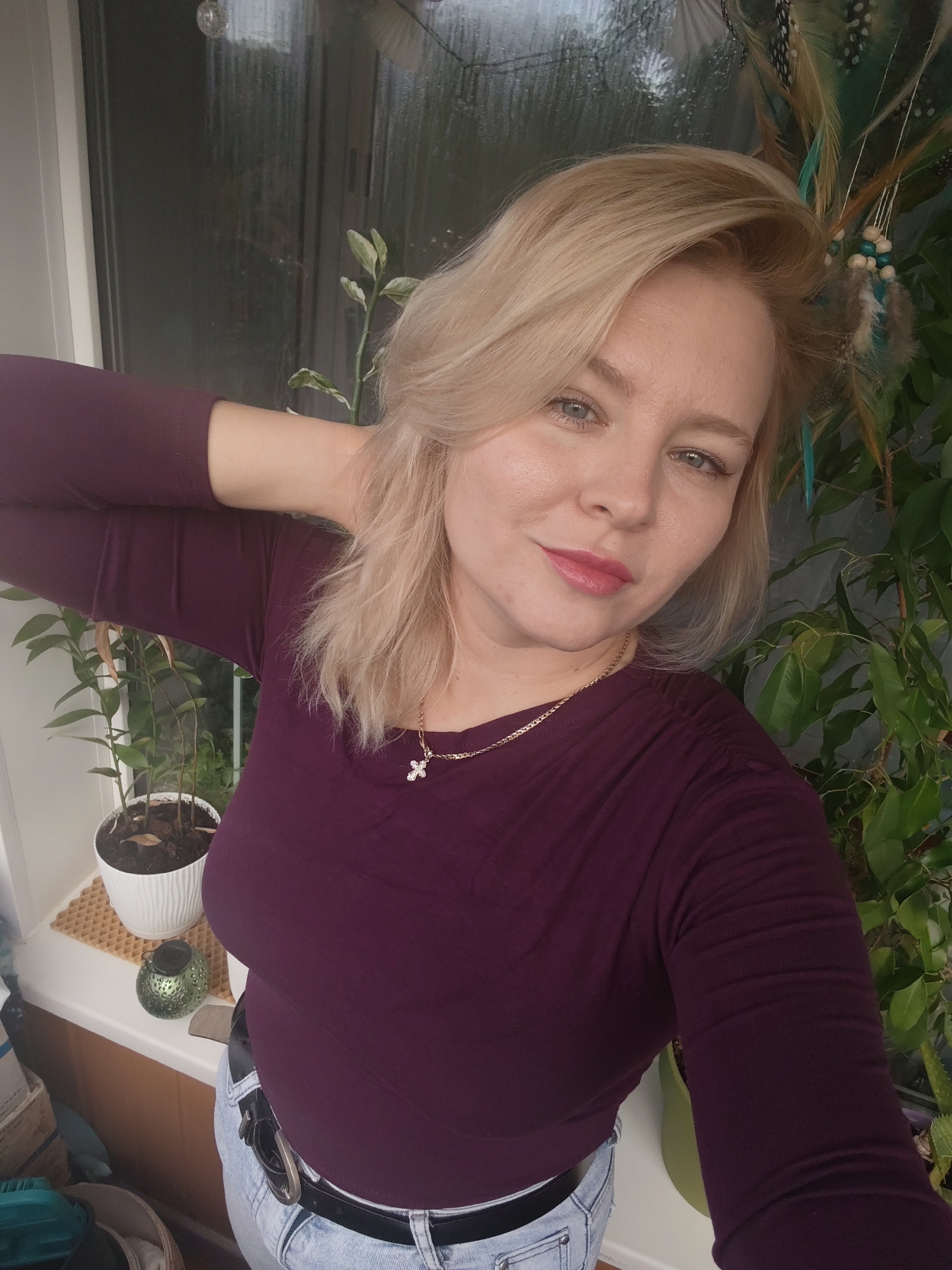 A blonde woman is captured in a selfie, her right arm resting on her head. She is wearing a plum-colored, long-sleeved shirt and light-wash denim jeans with a black belt. A gold necklace with a cross pendant adorns her neck. She is positioned indoors, with a window showing a blurred, rainy exterior behind her. To her left, a small potted plant sits on a windowsill. To her right, a large green plant with leaves and decorative feathers is visible. The overall setting suggests a residential interior, possibly in Izhevsk, Russia, given the context.